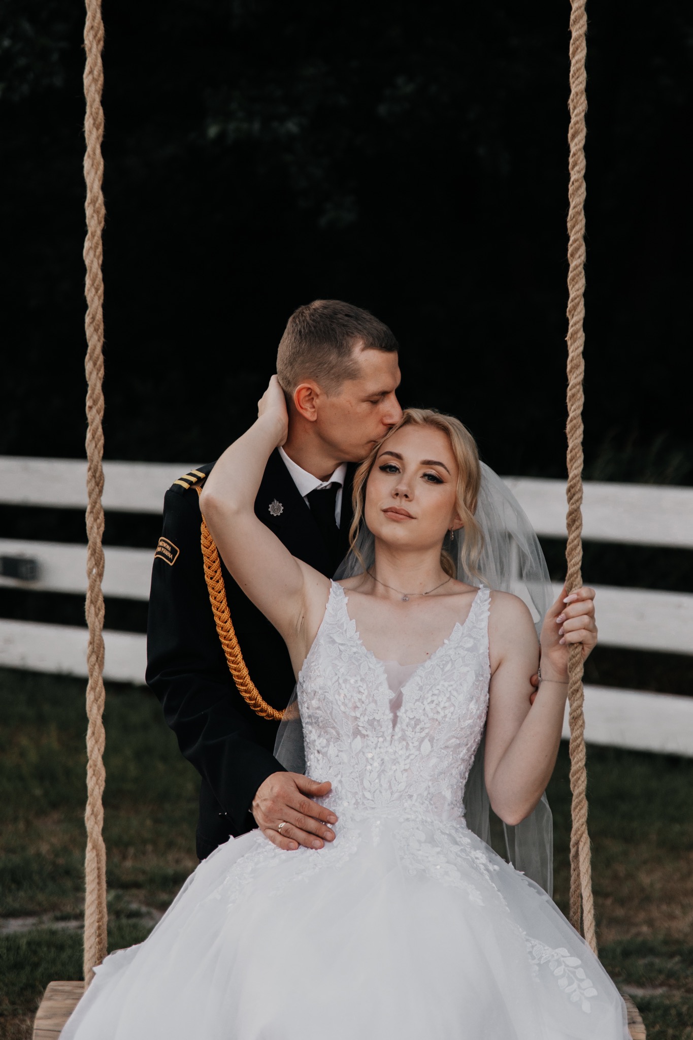Para młoda: panna młoda w koronkowej sukni i welonie, pan młody w mundurze, na huśtawce z linami, w tle białe ogrodzenie.