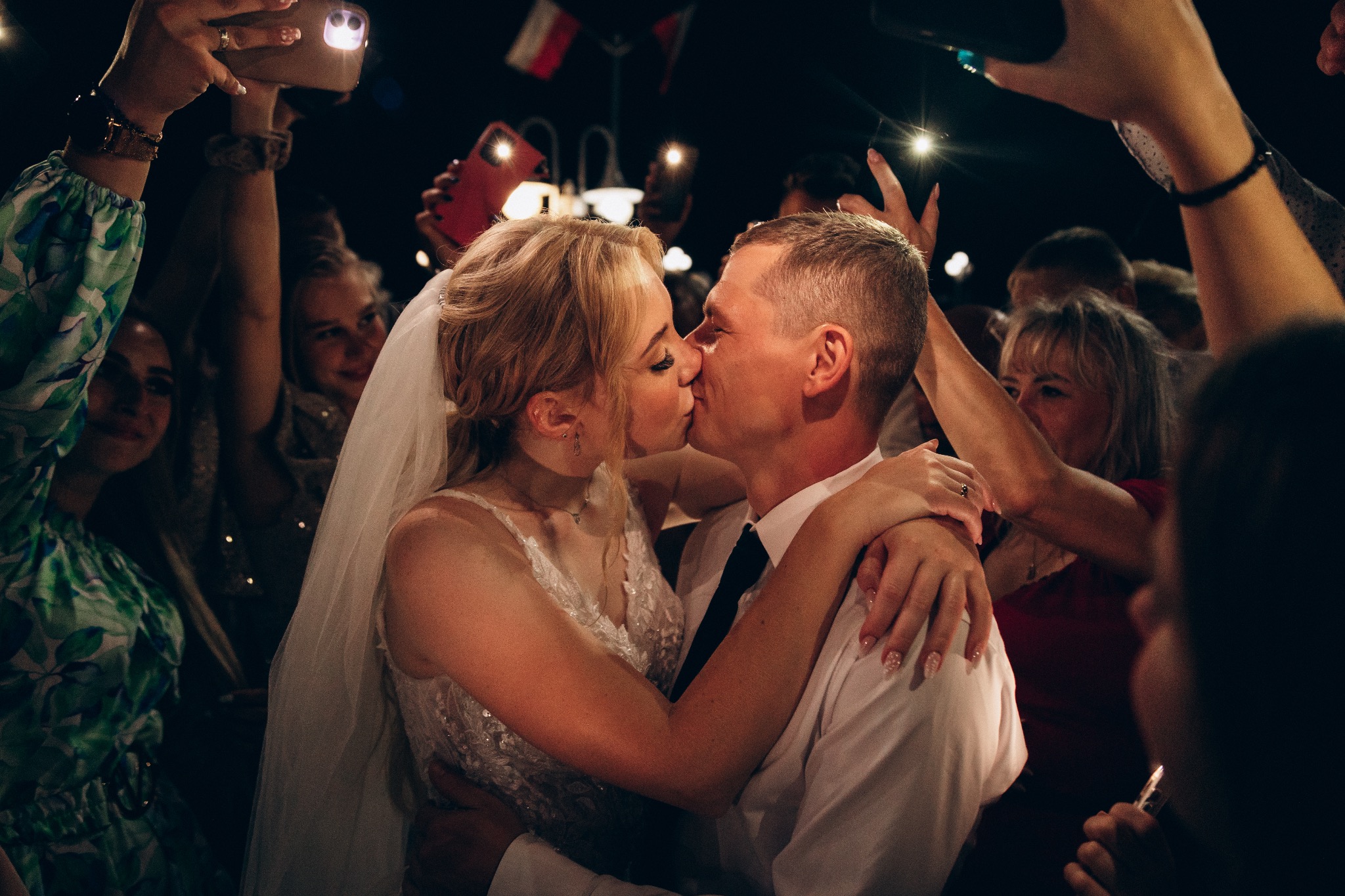 Pocałunek nowożeńców w otoczeniu gości weselnych z telefonami w górze, oświetlonych lampami błyskowymi.