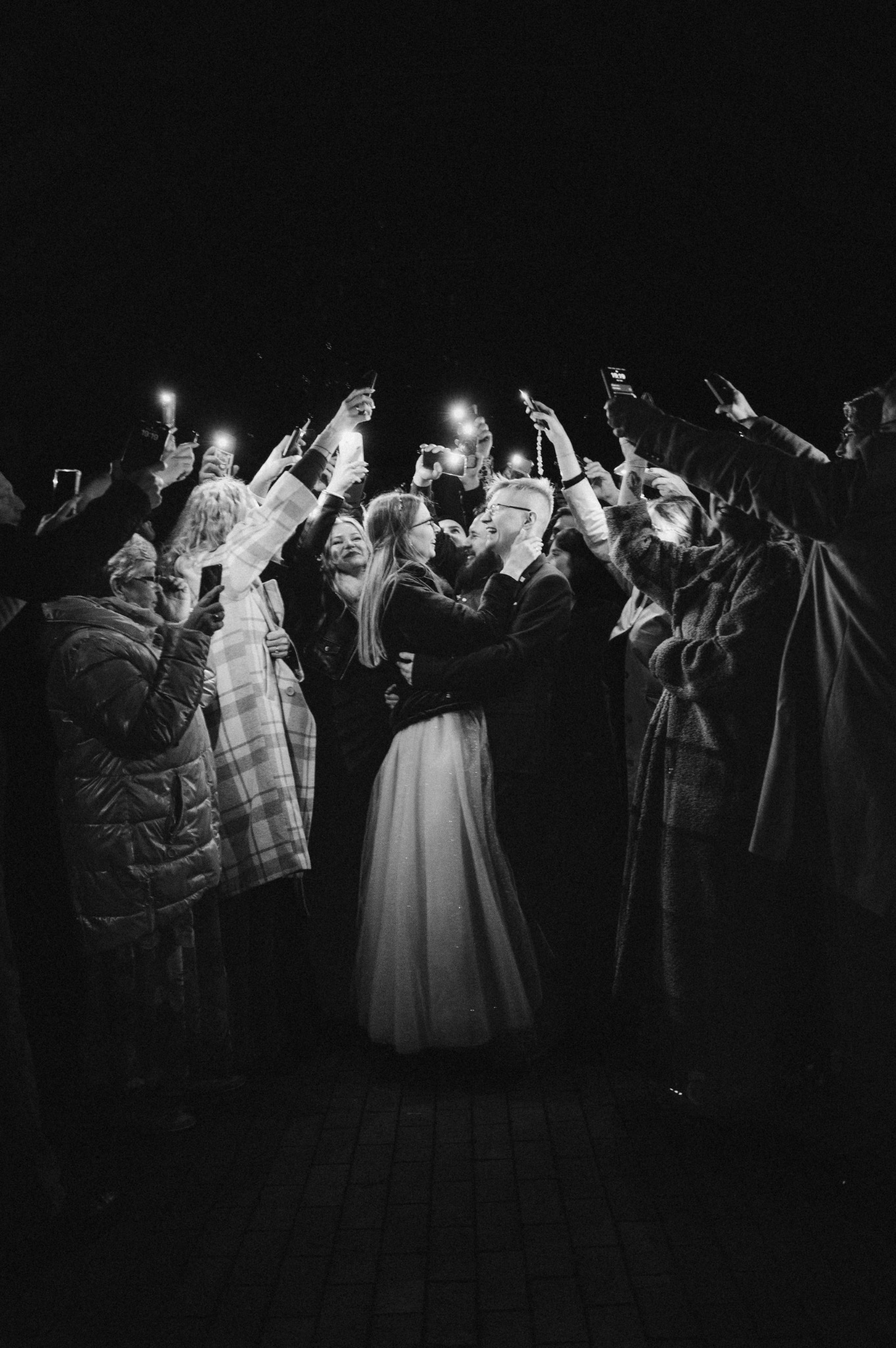 Czarno-białe zdjęcie nowożeńców otoczonych przez gości z telefonami w Nowej Soli. Para obejmuje się, a goście robią zdjęcia z uniesionymi telefonami.