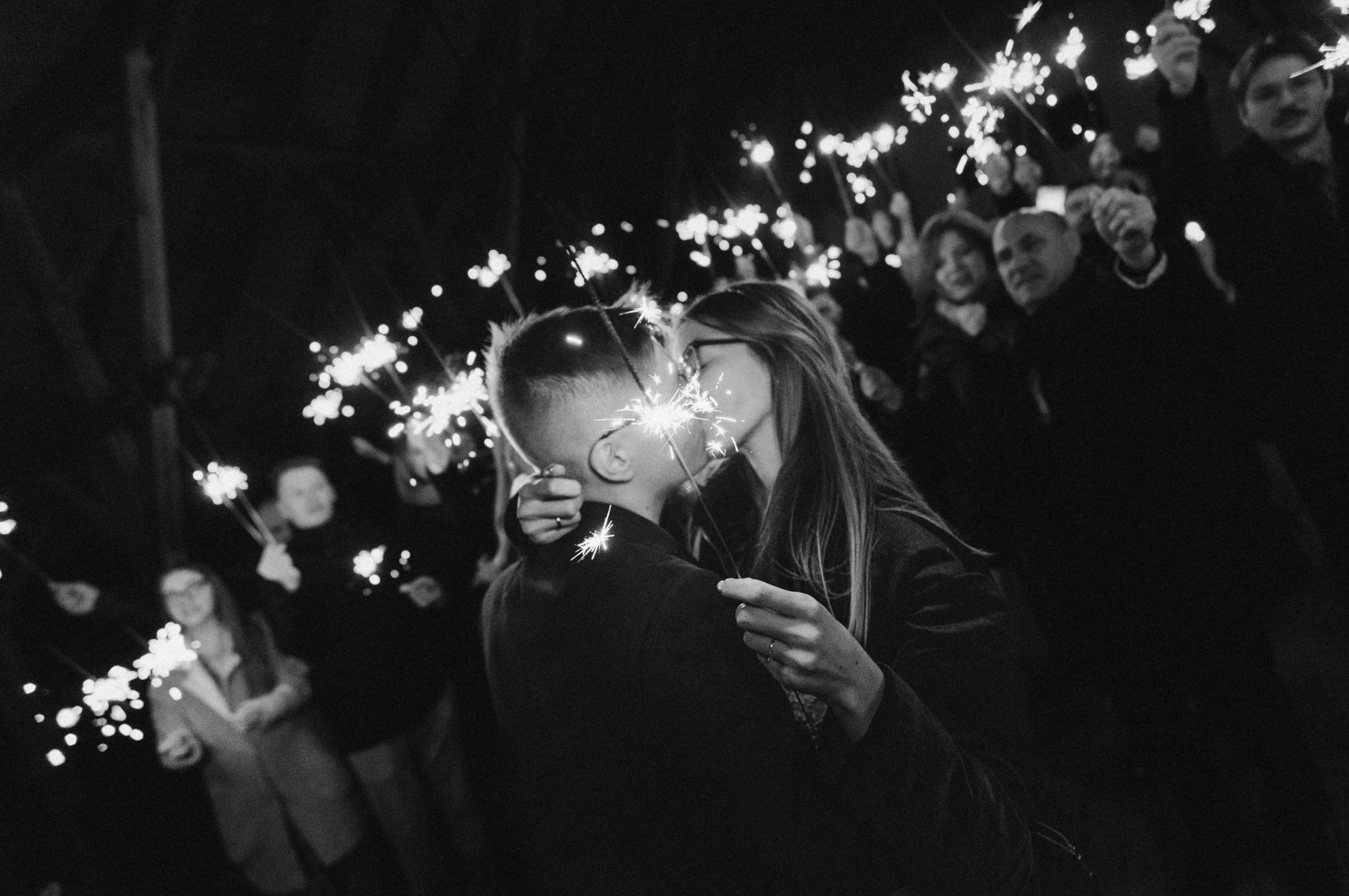 Czarno-białe zdjęcie całującej się pary młodej otoczonej gośćmi z zimnymi ogniami podczas wieczornego przyjęcia weselnego.
