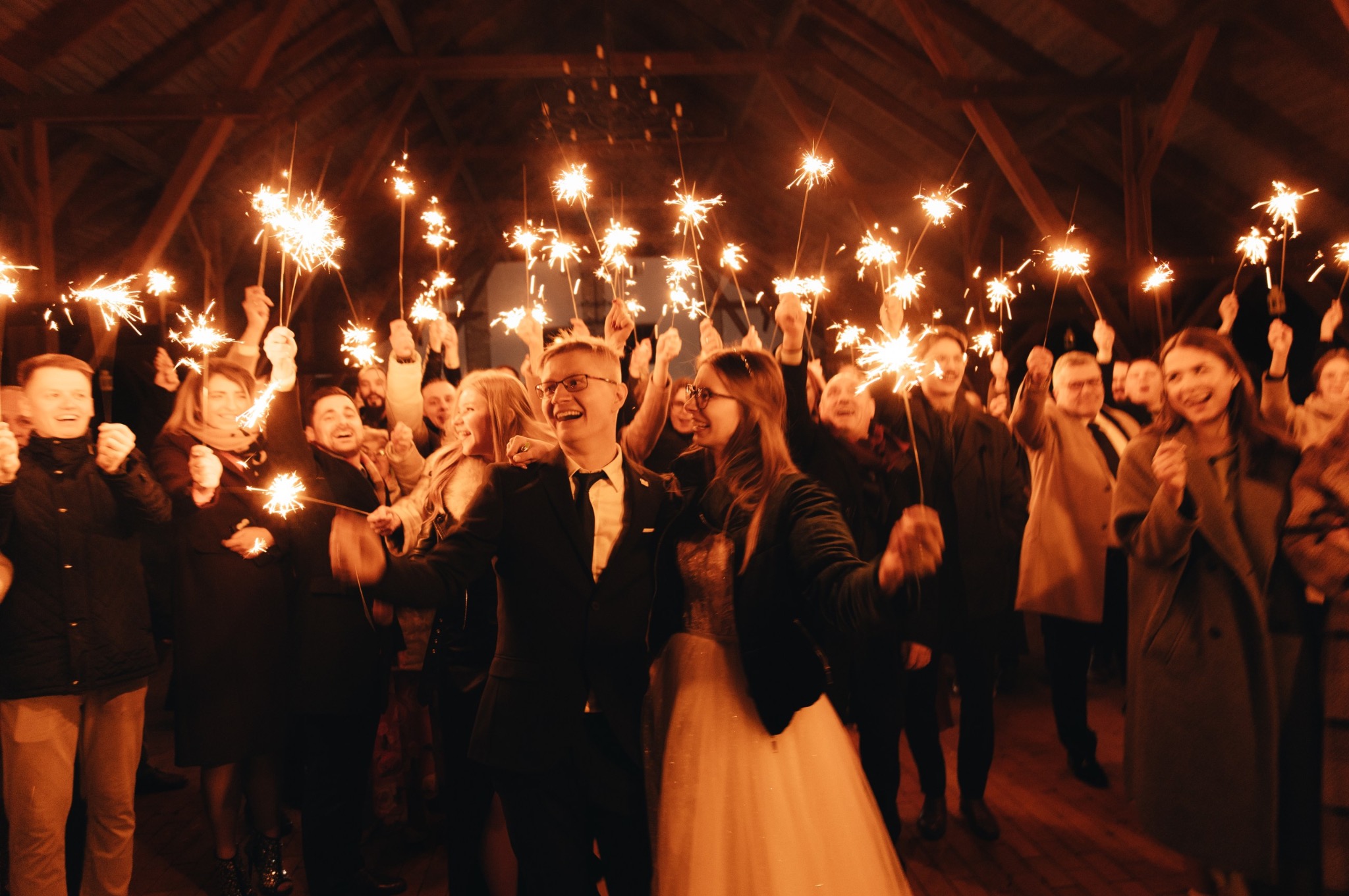 Uśmiechnięta para młoda otoczona gośćmi z zimnymi ogniami pod drewnianą konstrukcją dachu. Radosna atmosfera świętowania.