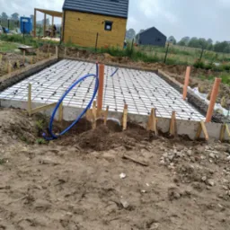 Przygotowanie płyty fundamentowej: zbrojenie stalowe na styropianie w szalunku, widoczne rury instalacyjne i budynek w tle.