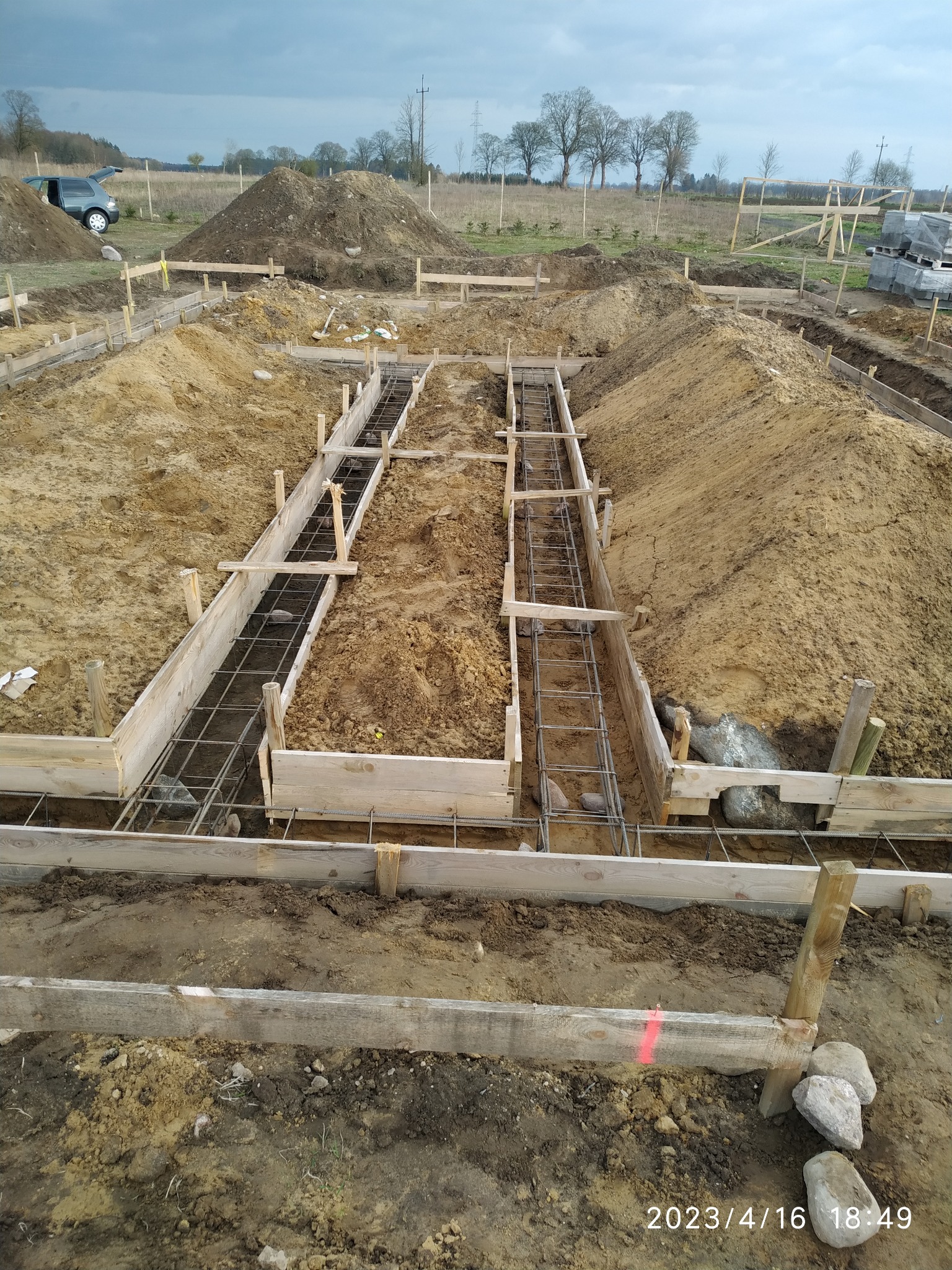 Przygotowanie wykopu pod fundamenty z drewnianym szalunkiem i zbrojeniem stalowym, widoczne pryzmy ziemi i samochód w tle.