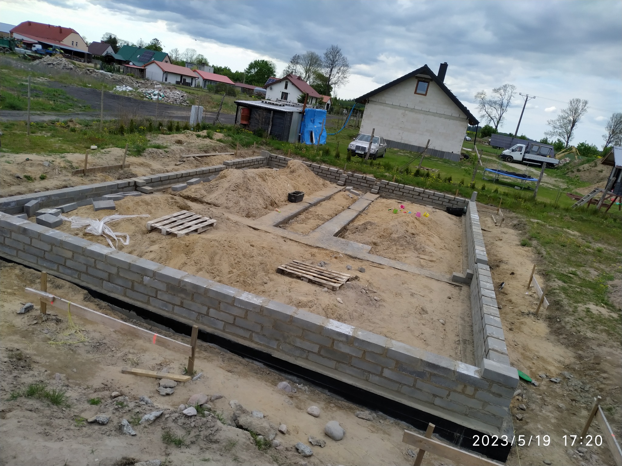 Wykop pod budowę domu z częściowo wzniesionymi ścianami fundamentowymi z bloczków betonowych, piasek w środku, drewniane palety, widoczne zbrojenie i izolacja przeciwwilgociowa.