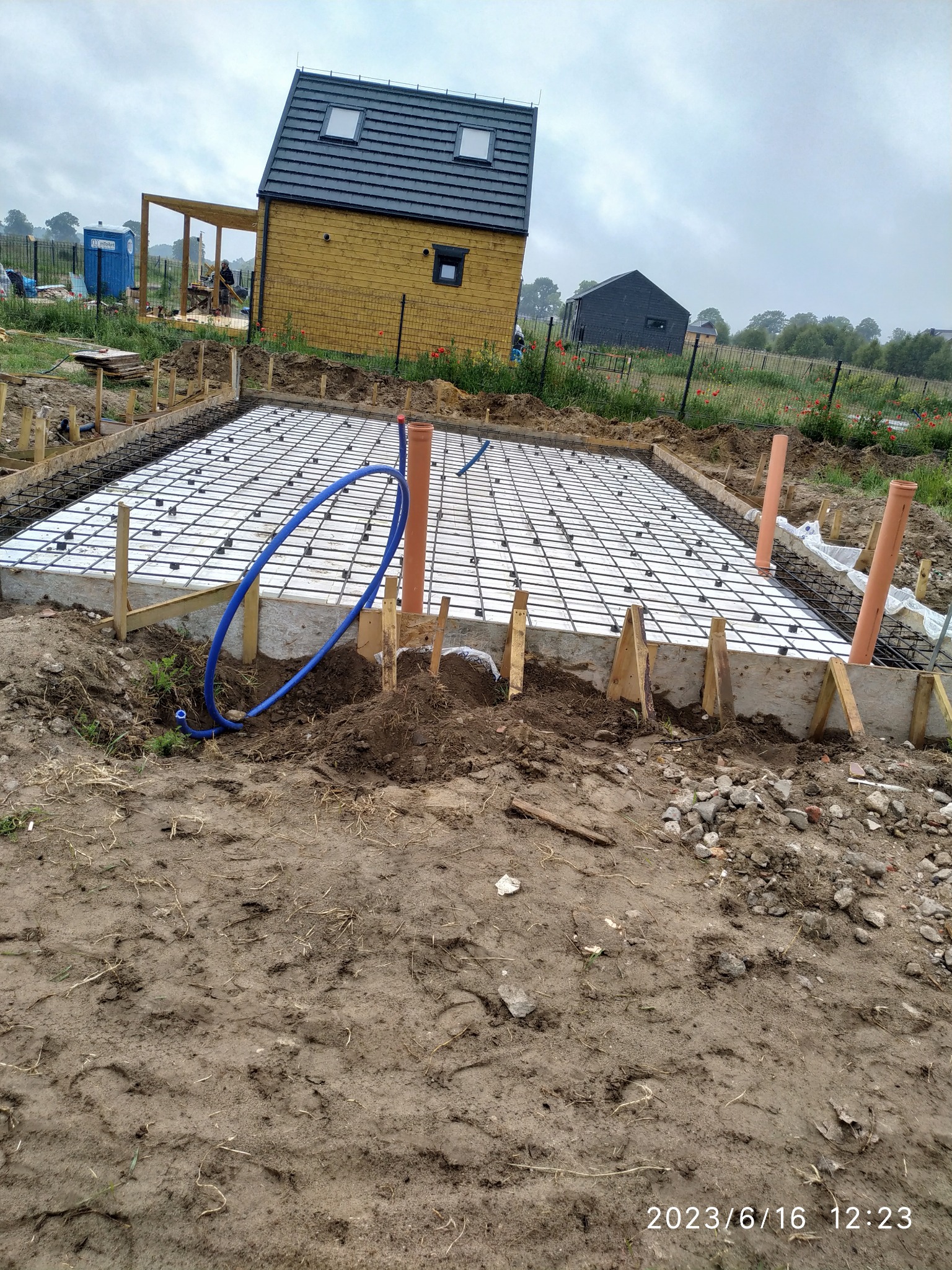 Przygotowanie płyty fundamentowej: zbrojenie stalowe na styropianie w szalunku, widoczne rury instalacyjne i budynek w tle.