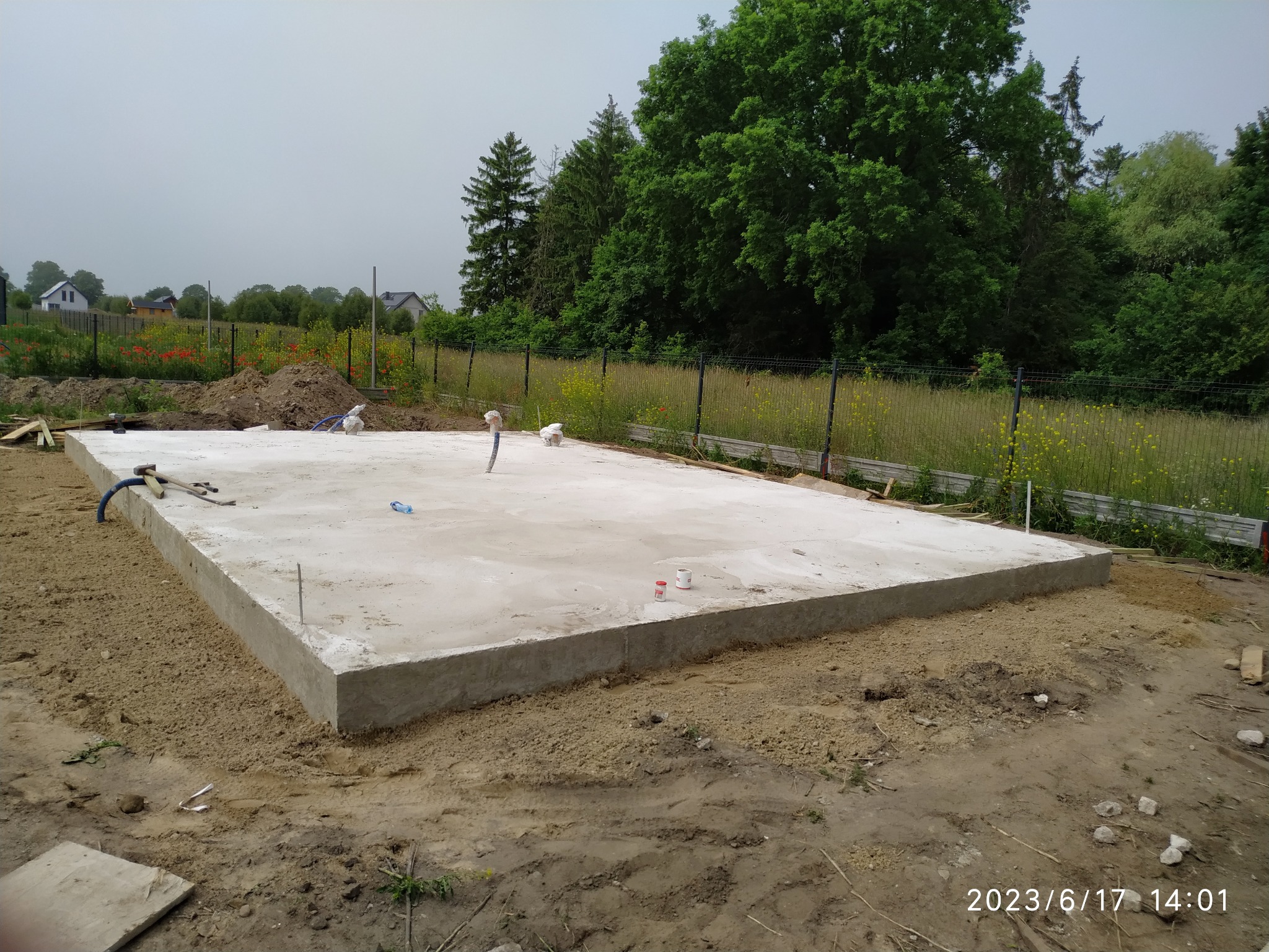 Wylany betonowy fundament płyty na gruncie, widoczne wystające rury instalacyjne, w tle ogród z kwitnącymi makami.