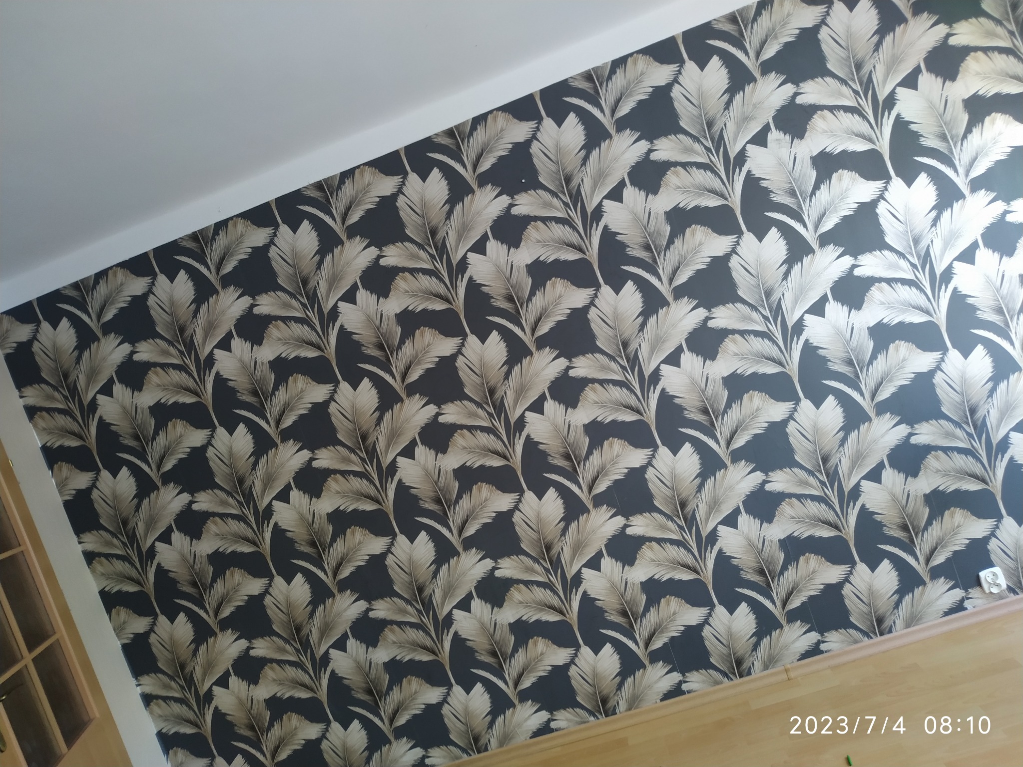Ściana pokryta tapetą w motywy liści palmowych w odcieniach szarości i beżu, widoczny fragment podłogi i sufitu, ujęcie z dołu.