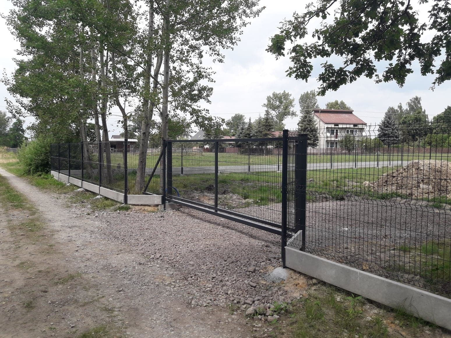 Czarna metalowa brama przesuwna z siatki na betonowej podmurówce, widok z zewnątrz na żwirowej drodze, w tle dom z czerwonym dachem i drzewa.