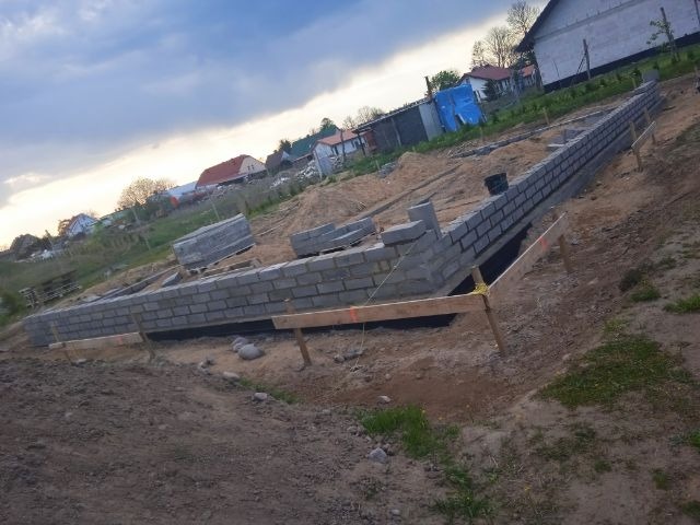 Wznoszenie fundamentów z bloczków betonowych na gruncie, widoczne drewniane szalunki i izolacja pionowa, w tle zabudowa jednorodzinna i pochmurne niebo.