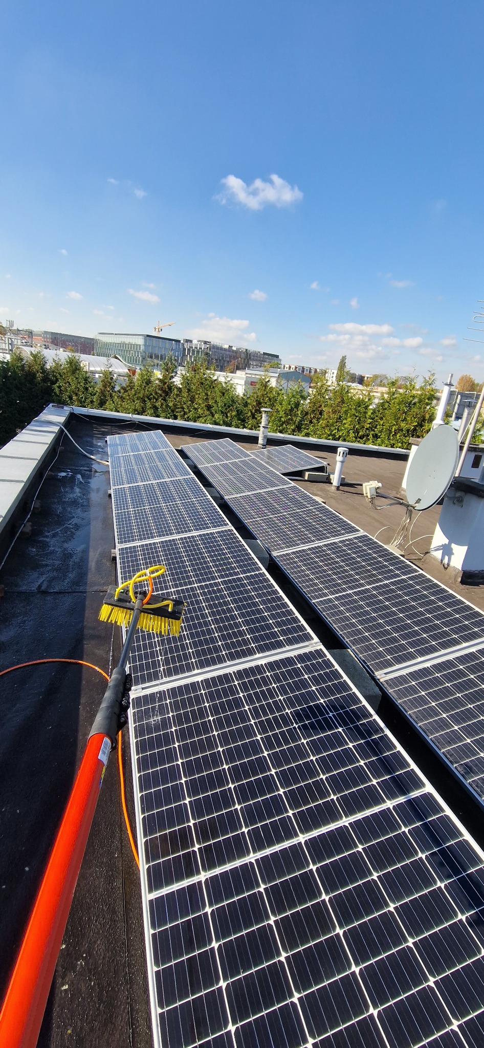 Czyszczenie paneli słonecznych na dachu płaskim za pomocą szczotki na wysięgniku, w tle budynki i niebieskie niebo.