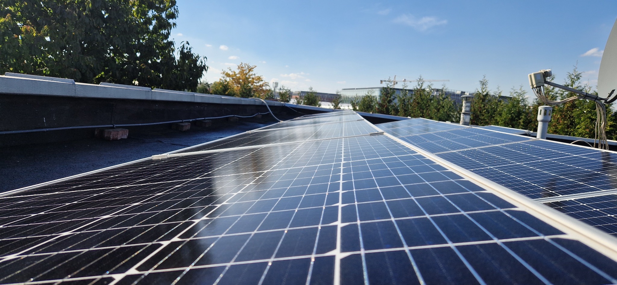 Widok z góry na panele słoneczne na płaskim dachu budynku z widocznymi przewodami, anteną satelitarną, kominami wentylacyjnymi i zielenią w tle.