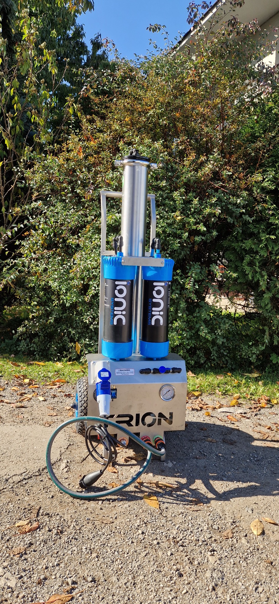 Mobilna stacja filtracyjna Ionic Systems na kółkach z dwoma pionowymi filtrami, widoczny manometr i podłączenia węży, na tle krzewów i niebieskiego nieba.