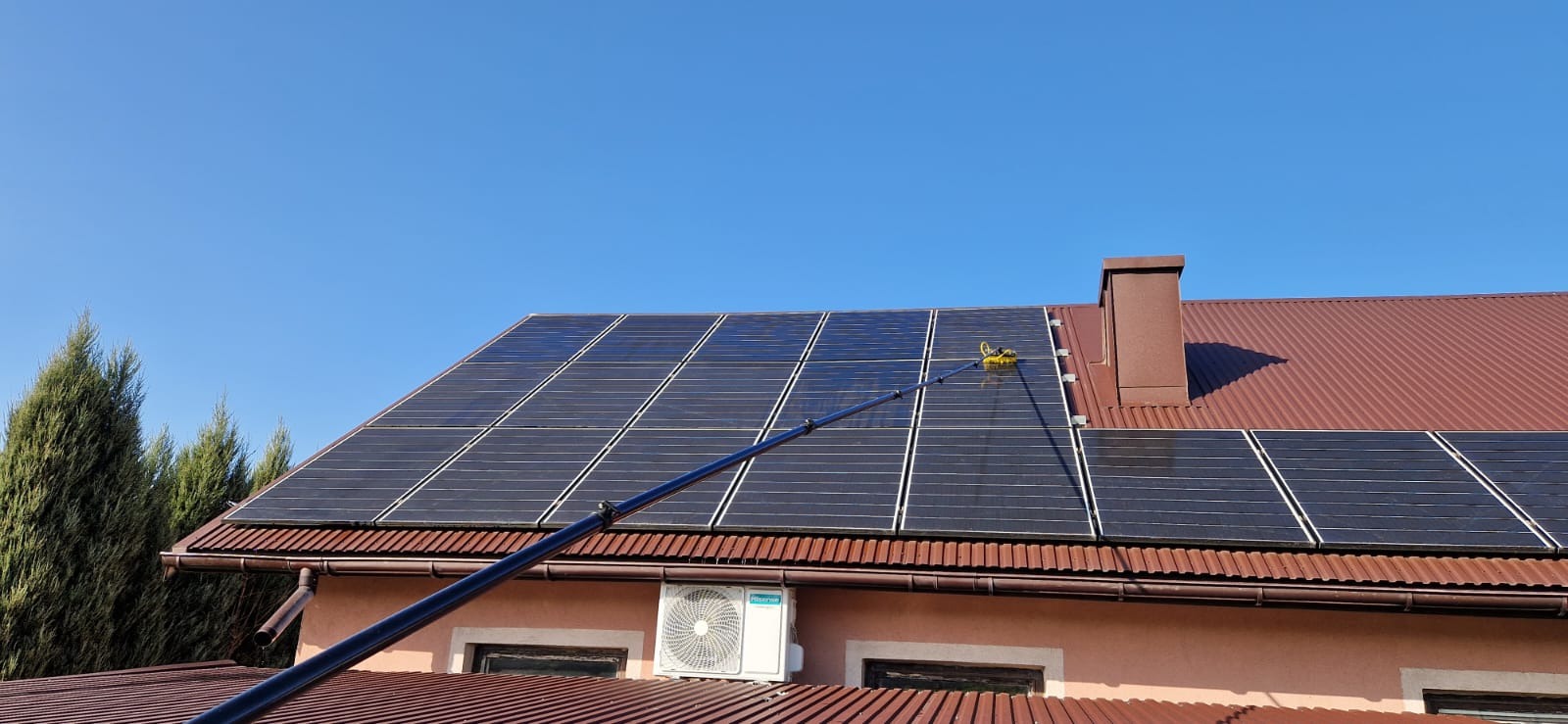 Czyszczenie paneli słonecznych na dachu domu jednorodzinnego przy użyciu teleskopowej lancy z żółtą szczotką, widoczny fragment dachu z brązową dachówką, komin, klimatyzator na ścianie i błękitne...
