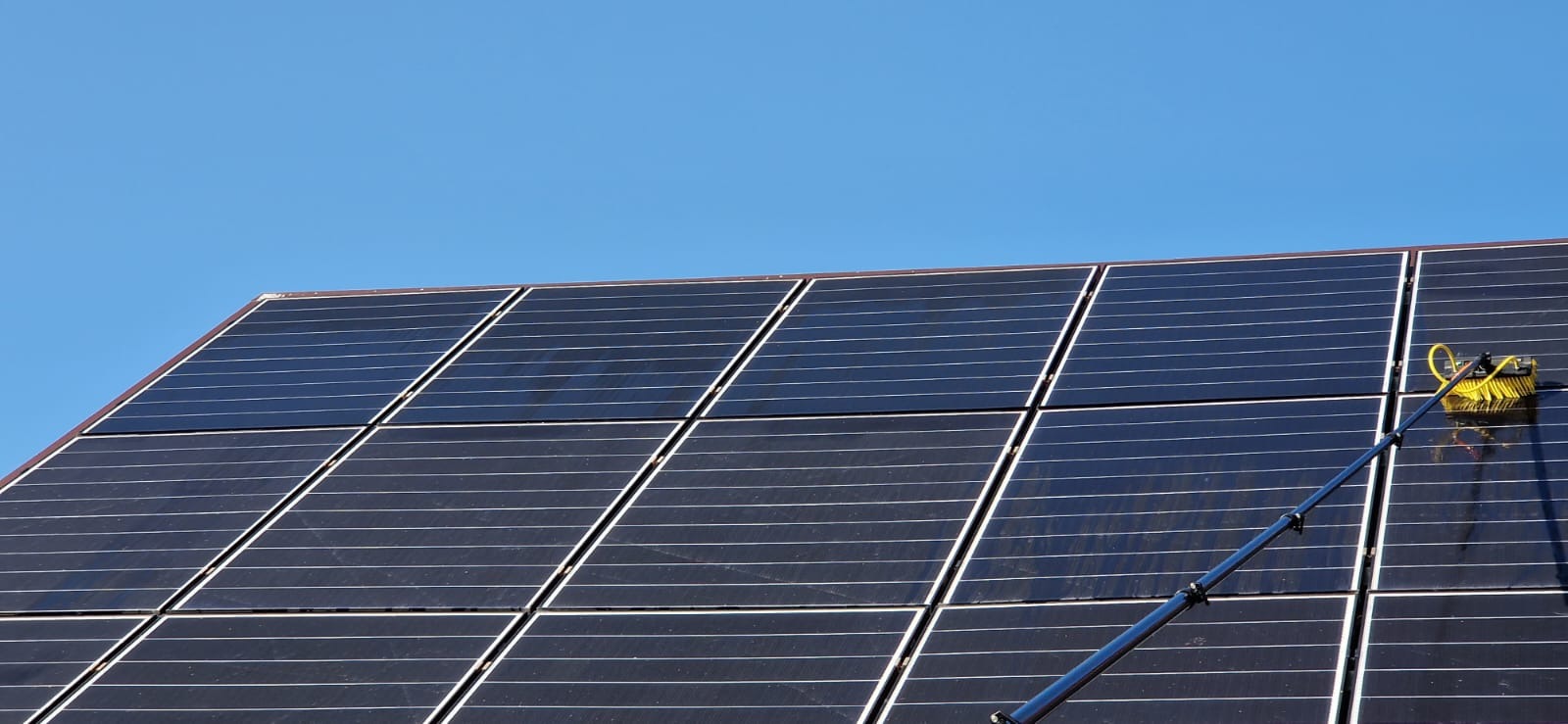 Czyszczenie paneli słonecznych na dachu za pomocą szczotki na teleskopowym kiju, błękitne niebo w tle.