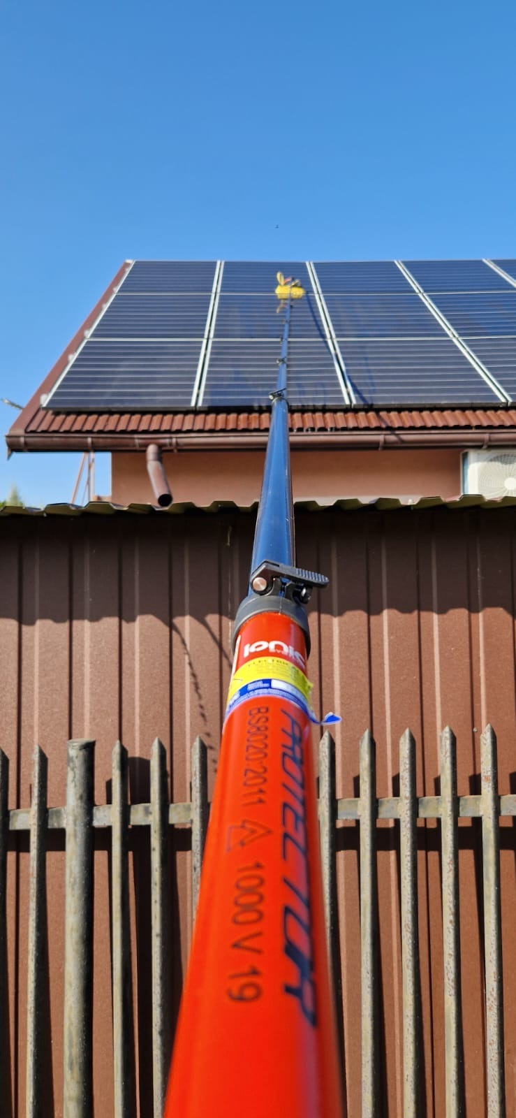 Czyszczenie paneli słonecznych na dachu domu za pomocą teleskopowej lancy z żółtą szczotką, widok od dołu na tle błękitnego nieba.
