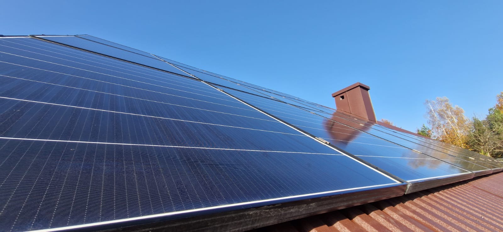 Panel słoneczny na dachu z brązowej blachy, widoczny komin i fragment drzew z żółtymi liśćmi na tle błękitnego nieba.