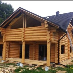 Taras Samotii STV - Surowy drewniany dom z bali w trakcie budowy, z widoczną konstrukcją dachu i oknami bez ram, drabina oparta o ścianę, teren wokół budynku częściowo zagospodarowany.