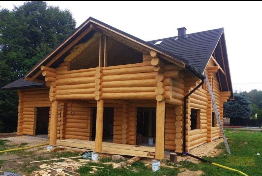 Surowy drewniany dom z bali w trakcie budowy, z widoczną konstrukcją dachu i oknami bez ram, drabina oparta o ścianę, teren wokół budynku częściowo zagospodarowany.