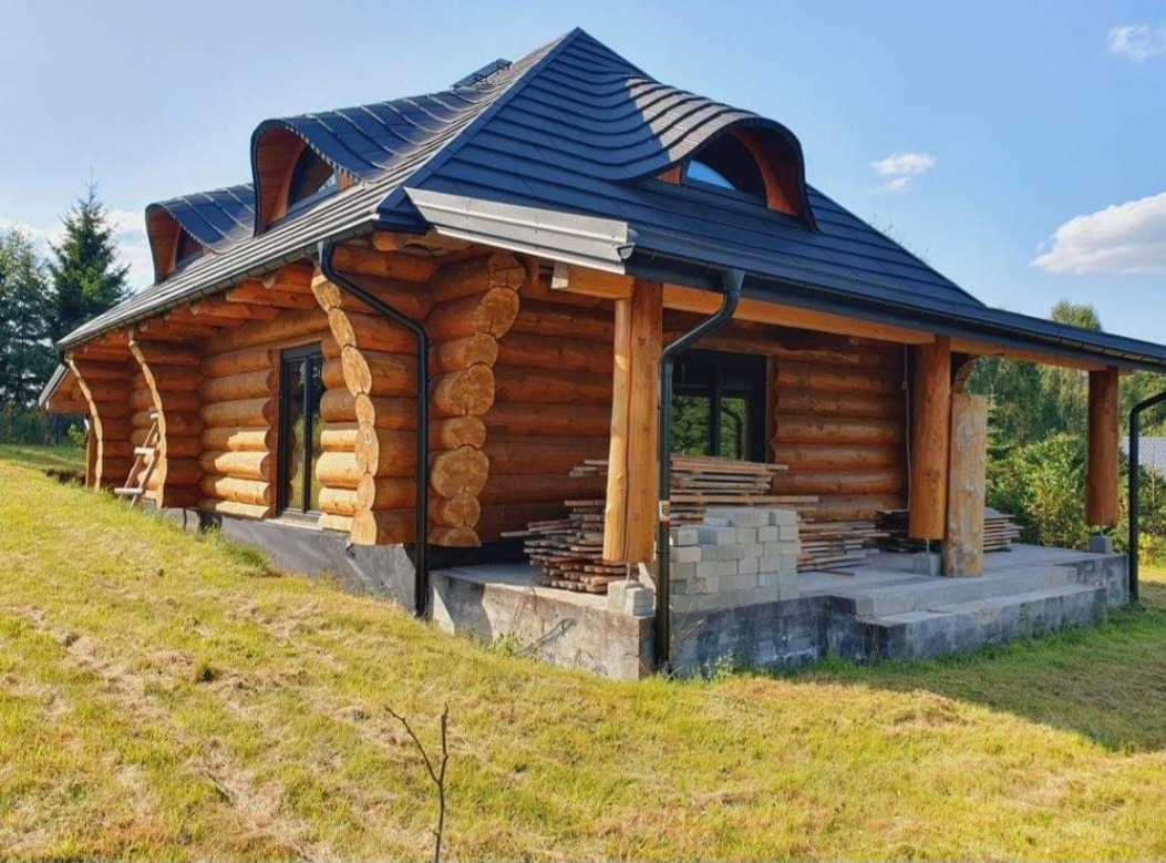 Nowoczesny dom z bali z oryginalnym dachem krytym czarną dachówką, widoczne przygotowane deski i bloczki betonowe na tarasie.