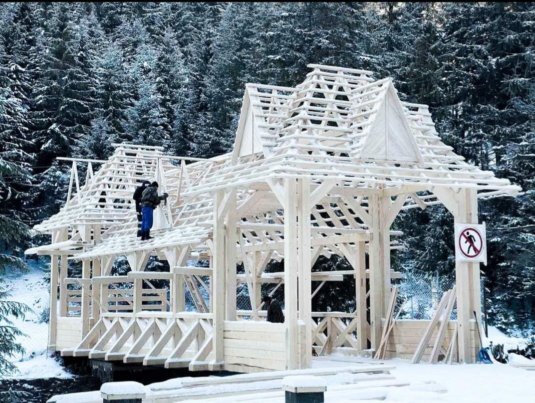 Budowa drewnianej altany w zimowym krajobrazie leśnym, widoczne elementy konstrukcyjne dachu i pracujący na nim robotnicy, tabliczka zakazu przejścia w tle.