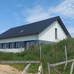 In Dom - Nowoczesny dom jednorodzinny z ciemnoszarą elewacją w dolnej części i białą w górnej, z ciemną dachówką, widziany zza drewnianego płotu i wysokiej trawy, obok widać budynek w trakcie budowy.