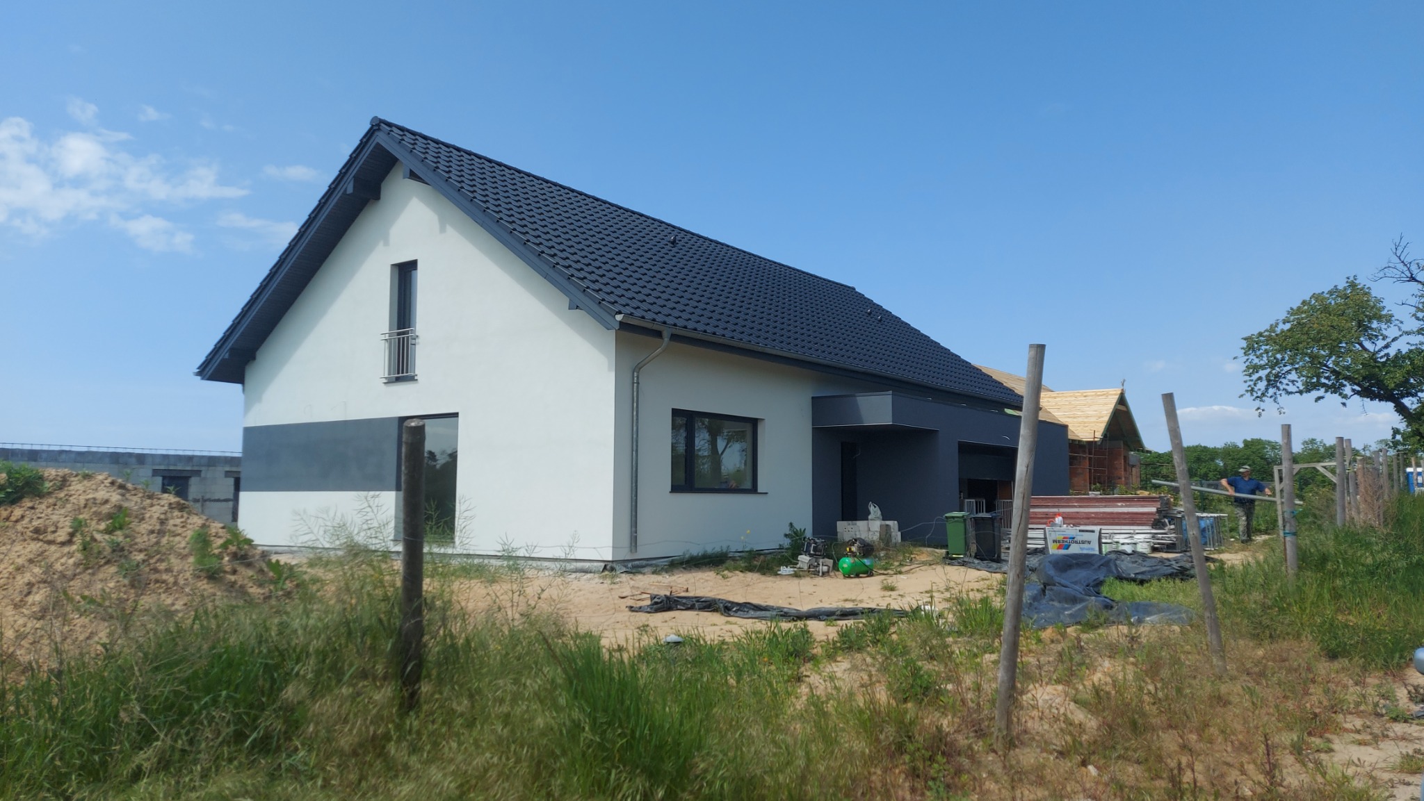 Nowoczesny dom jednorodzinny z szarą elewacją i ciemną dachówką, widoczny w trakcie budowy, z materiałami budowlanymi i częściowo wykończonym dachem sąsiedniego budynku w tle.