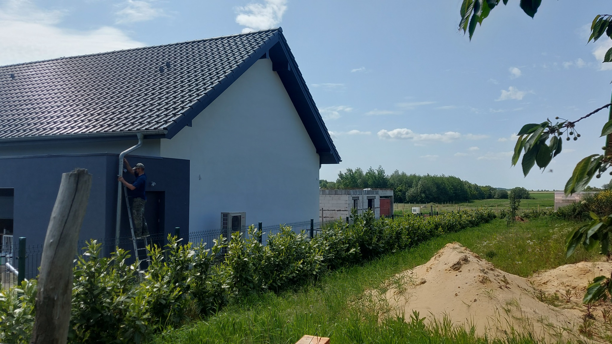 Pracownik na drabinie przy nowym domu z ciemną dachówką, montujący rynnę spustową, widok z zewnątrz na tle zieleni i błękitnego nieba.