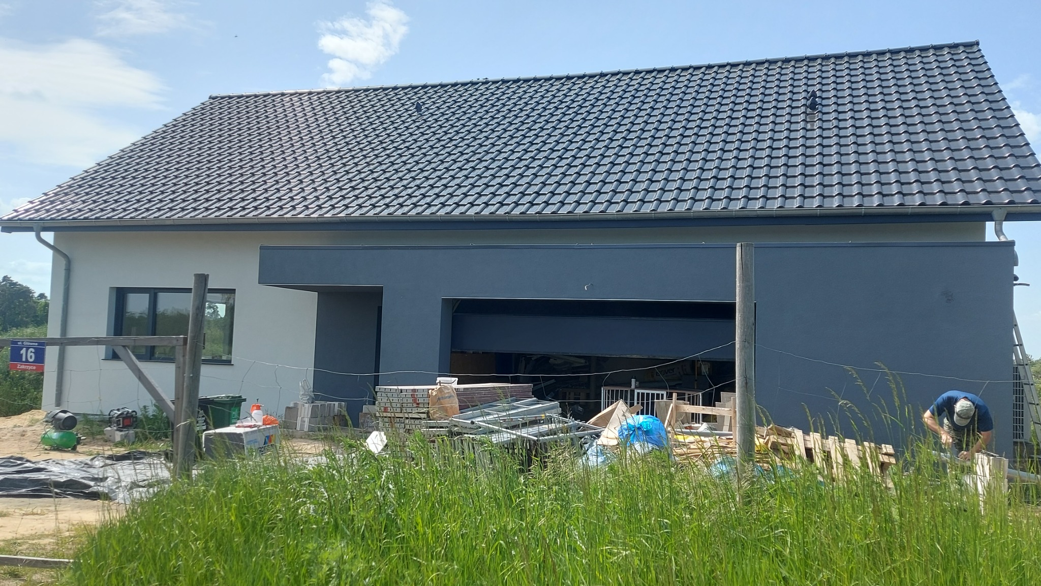 Nowoczesny dom jednorodzinny z grafitową elewacją i ciemnoszarą dachówką, widok zza wysokiej trawy, w tle pracownik przy pracy na zewnątrz.
