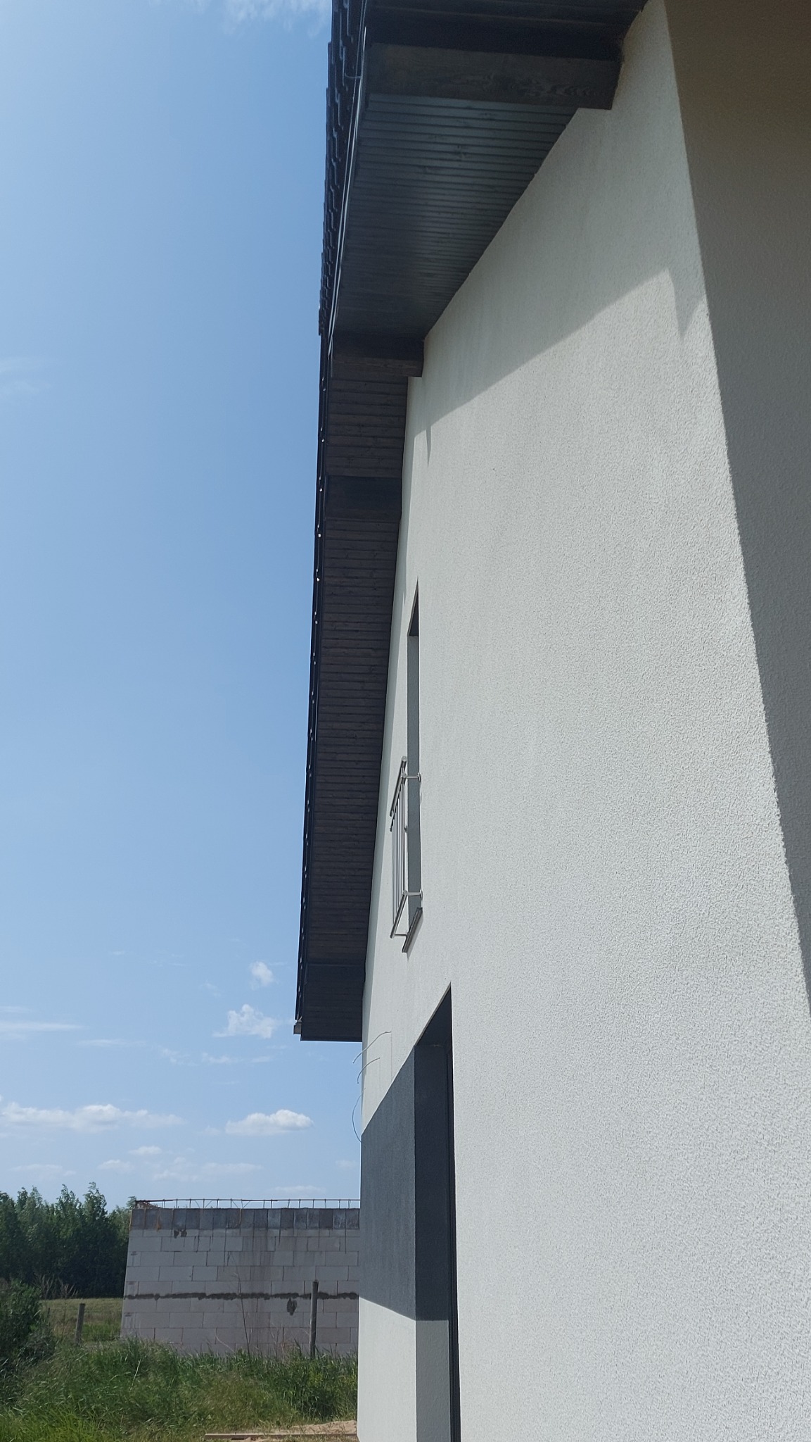 Jasna elewacja budynku z widoczną strukturą tynku, ciemne obramowanie okna i fragment dachu, w tle niezakończona budowa z bloczków betonowych.