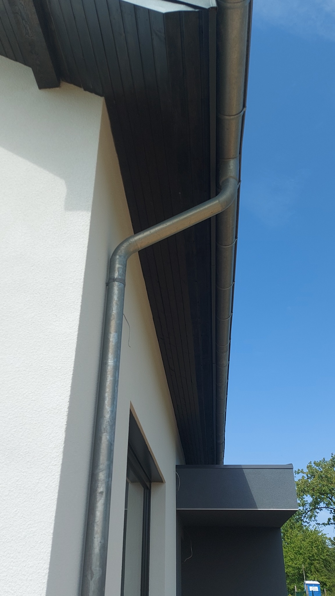 Nowa, ocynkowana rynna spustowa, zamontowana przy białej elewacji budynku z ciemnym, drewnianym podbitkiem dachowym i fragmentem okna.