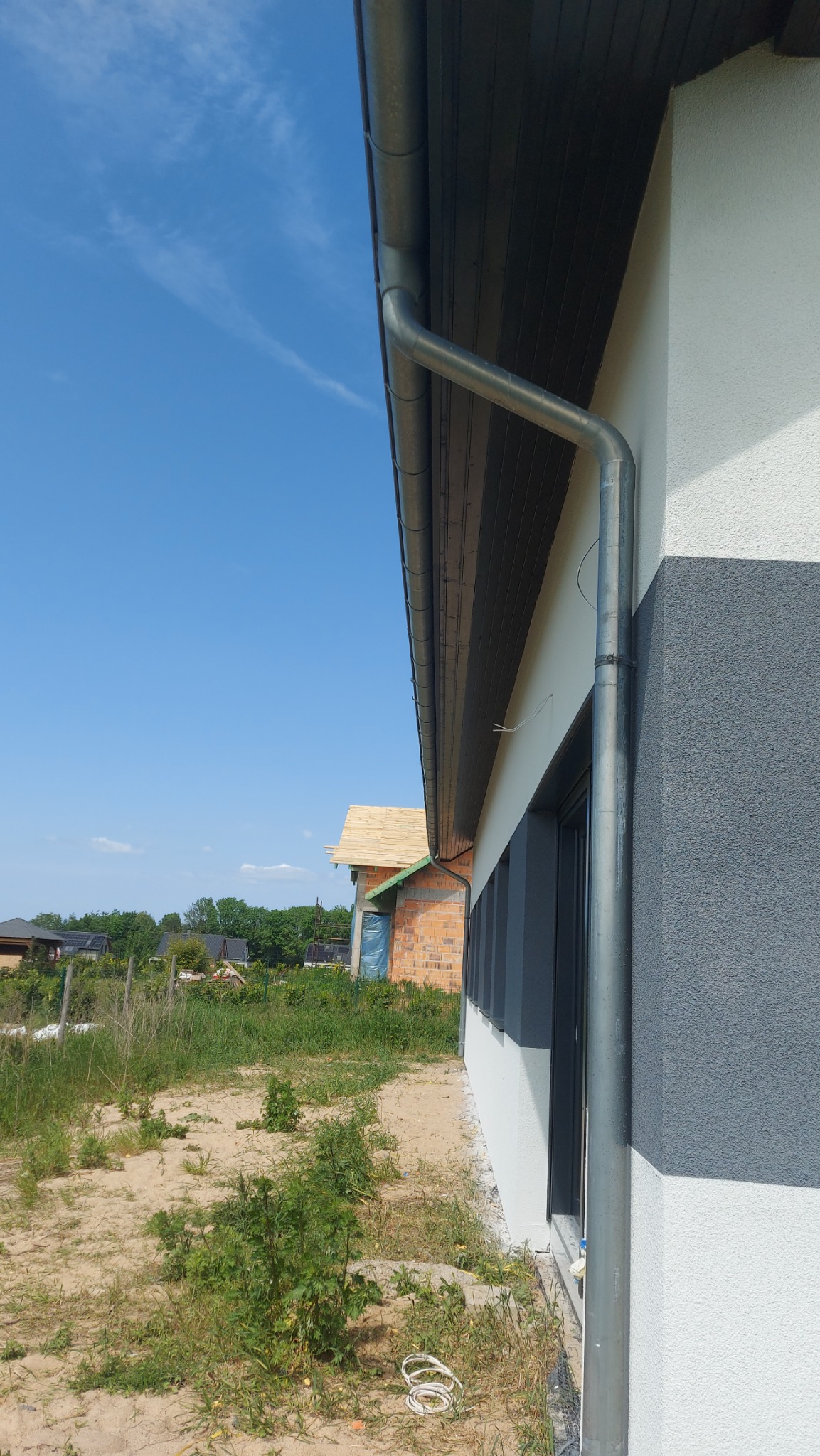 Szara metalowa rynna spustowa zamontowana na elewacji budynku z widocznym fragmentem dachu i elewacją w dwóch odcieniach szarości, w tle budynek w trakcie budowy.