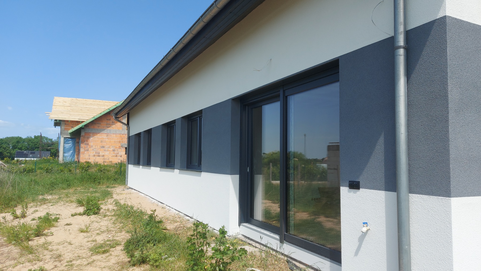 Dwukolorowa elewacja budynku jednorodzinnego z dużym oknem przesuwnym, widoczna rynna i fragment budynku w trakcie budowy w tle.
