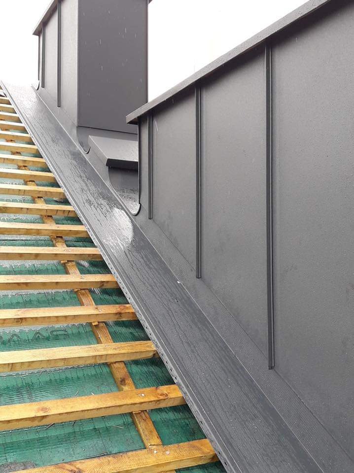 Szara, metalowa obudowa komina i fragment dachu z drewnianą konstrukcją, widoczne krople wody na metalowej powierzchni.