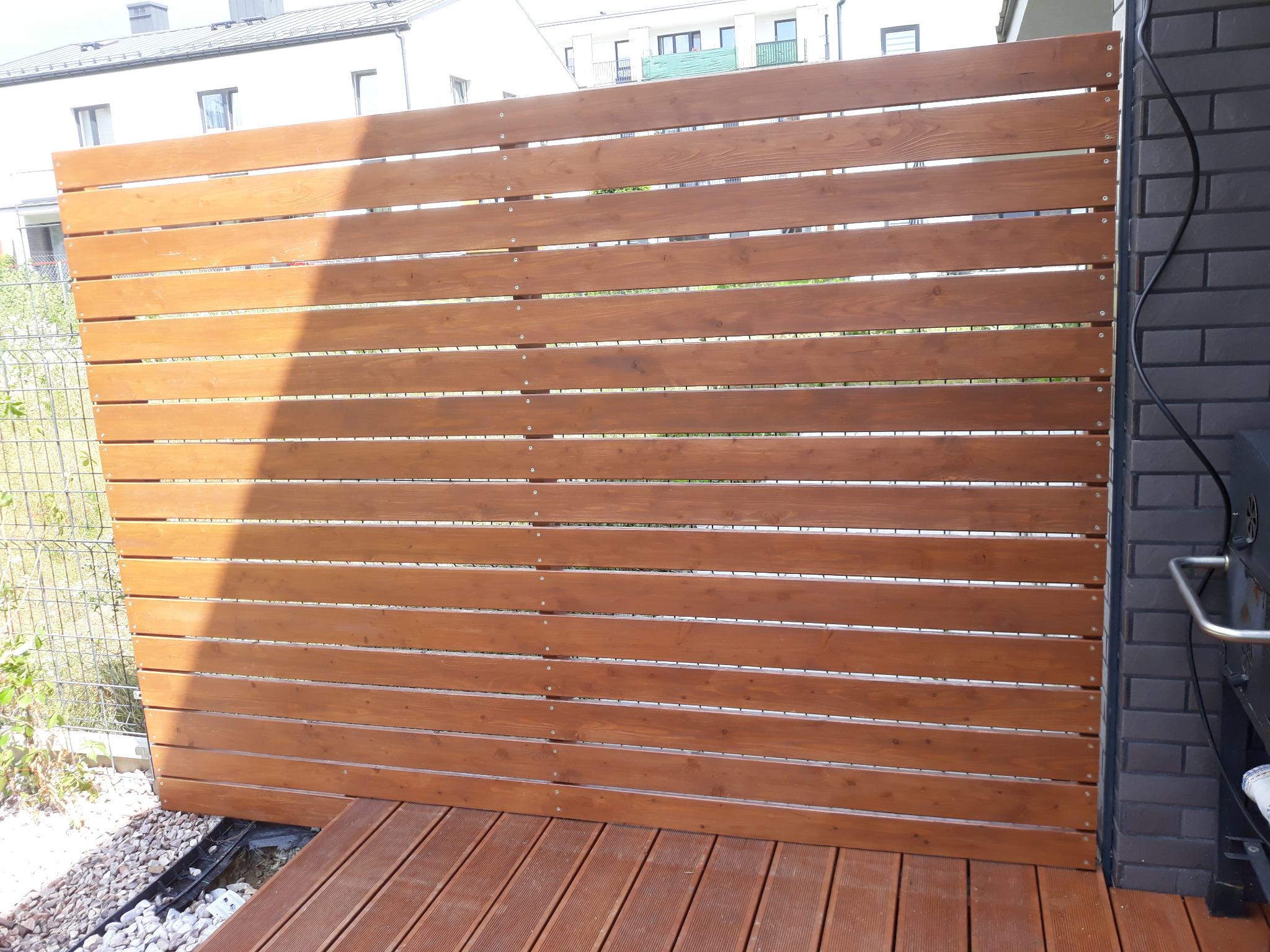 Drewniana przesłona z poziomych desek, mocowana śrubami, oddzielająca taras od ogrodu, z widoczną konstrukcją tarasu i fragmentem grilla.