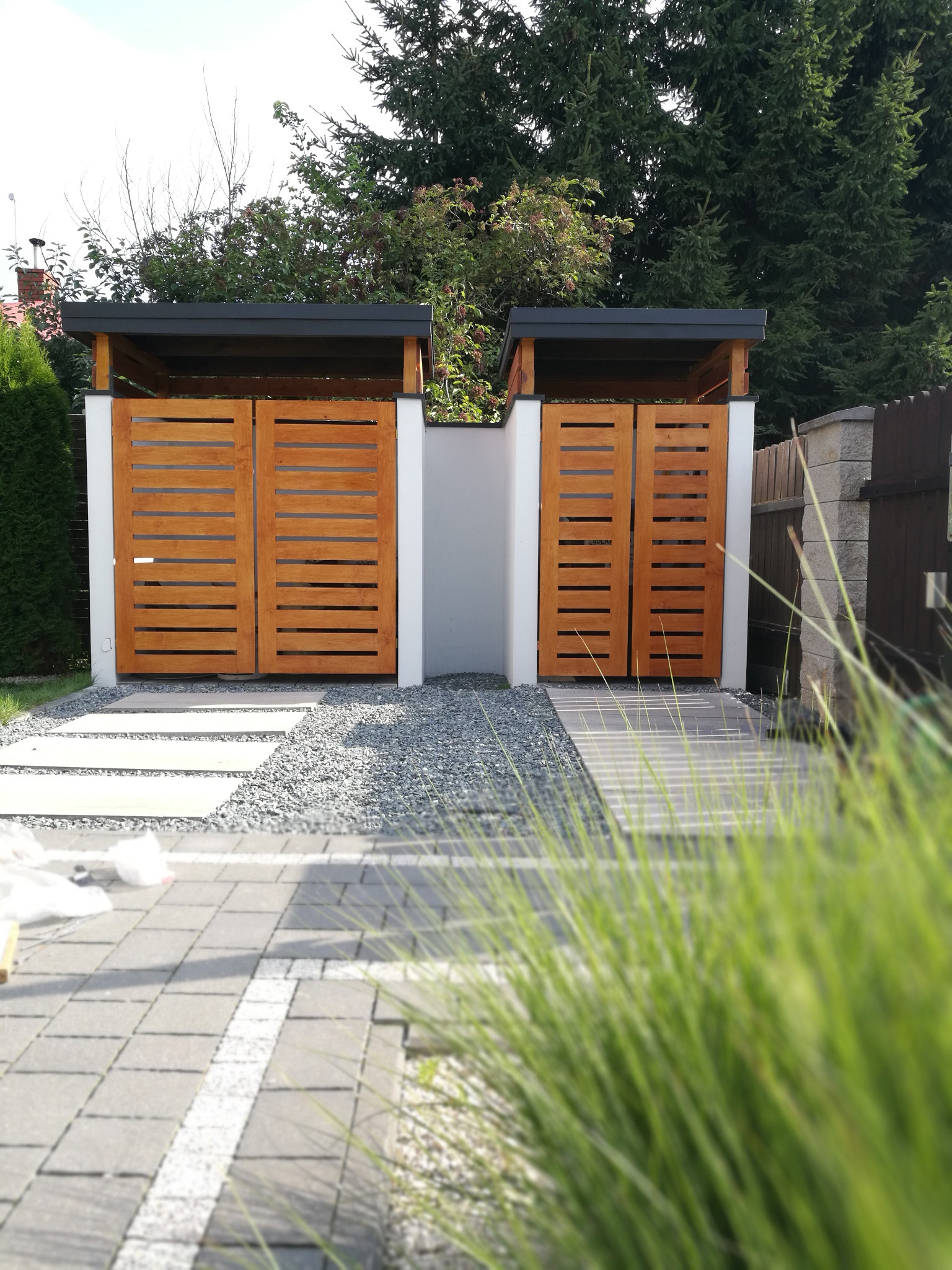 Dwa zadaszone pojemniki na śmieci z drewnianymi, poziomymi panelami, oddzielone białym murem, otoczone szarą żwirową nawierzchnią i betonowymi płytami chodnikowymi.