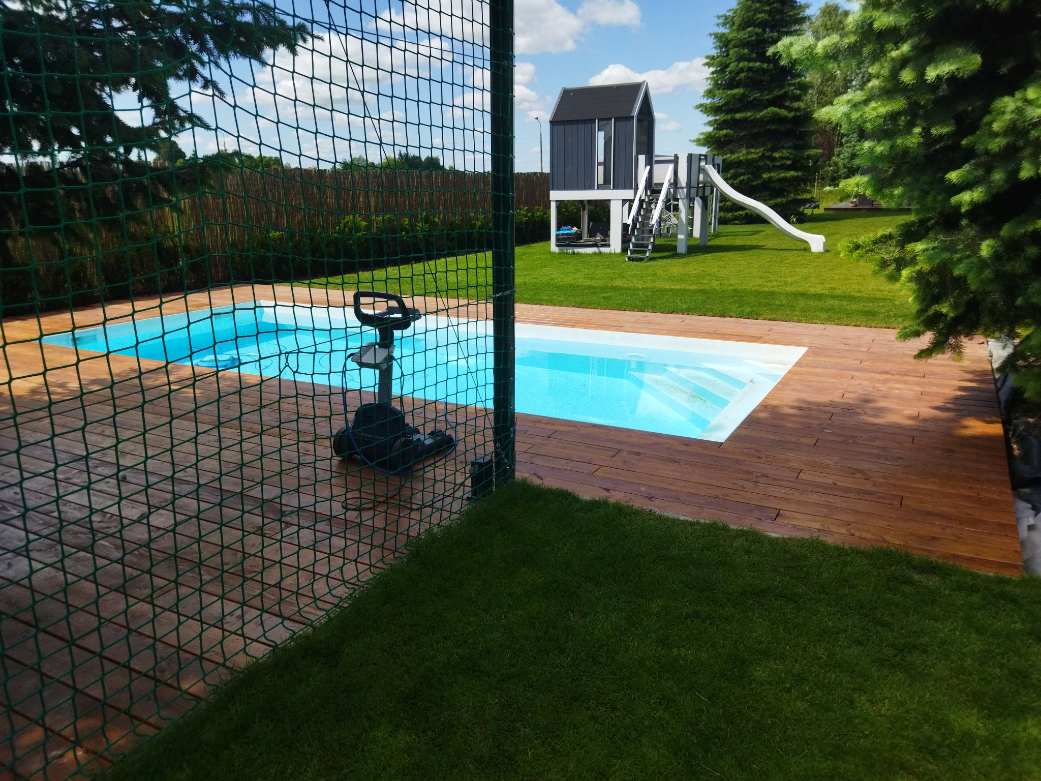 Nowoczesny basen z drewnianym tarasem i siatką zabezpieczającą, na tle zielonego trawnika z domkiem zabaw dla dzieci.