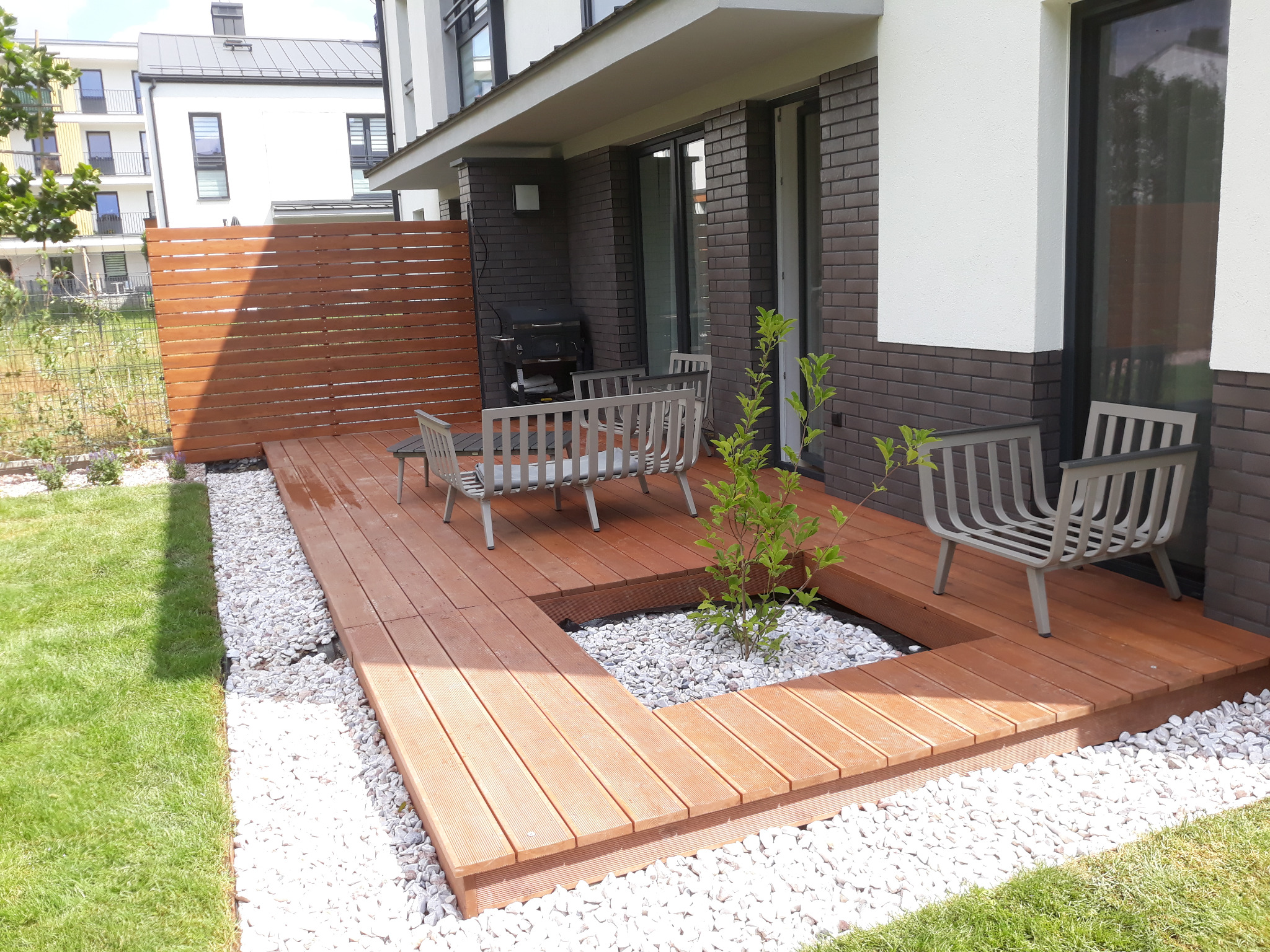 Nowoczesny taras drewniany z wyciętym miejscem na roślinę, otoczony białymi kamieniami, z metalowymi krzesłami i drewnianym ogrodzeniem w tle.