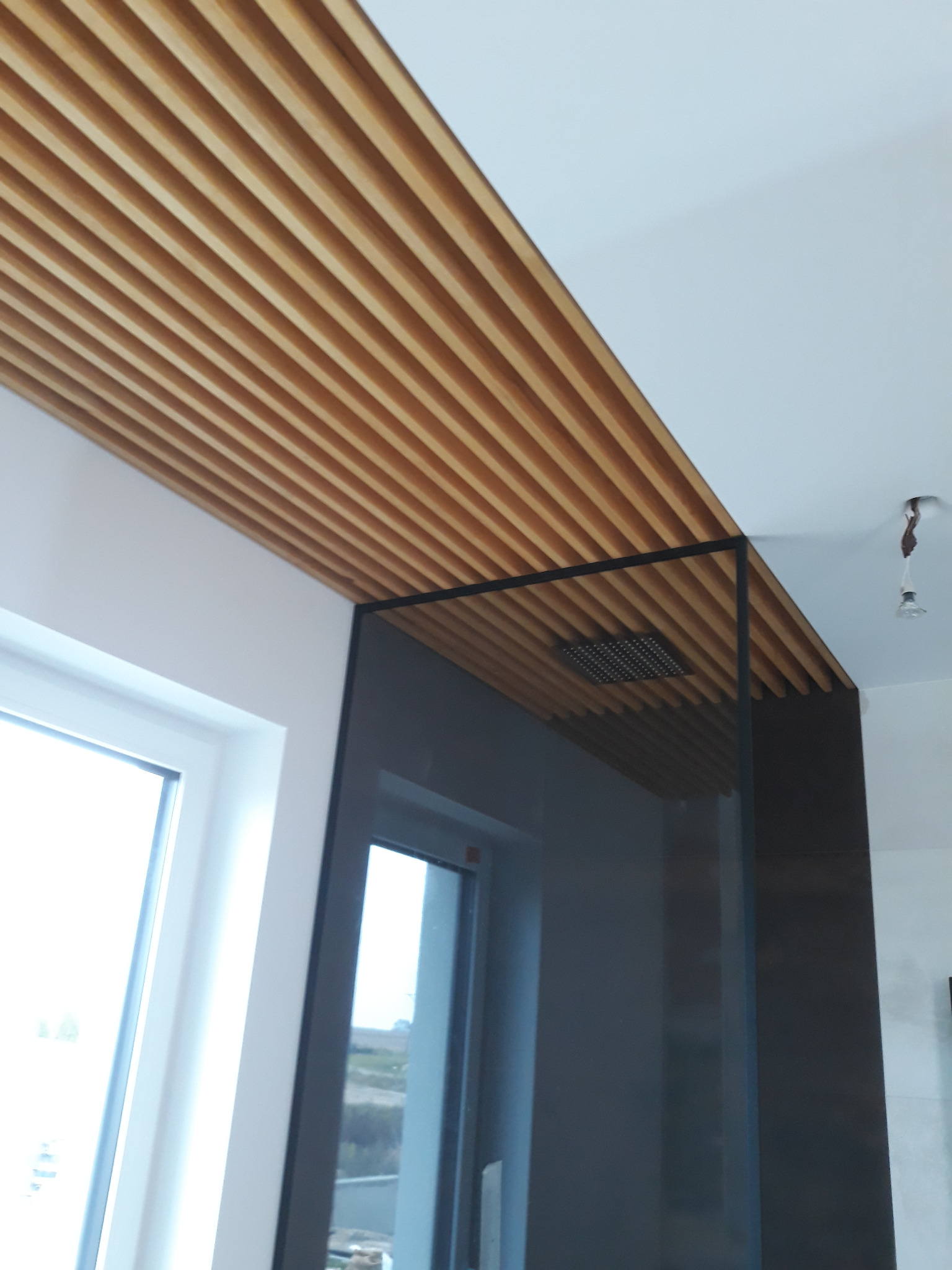 Drewniane lamele na suficie łazienki, zintegrowane z kabiną prysznicową z czarnymi profilami i deszczownicą, widok z dołu, okno z widokiem na zewnątrz.