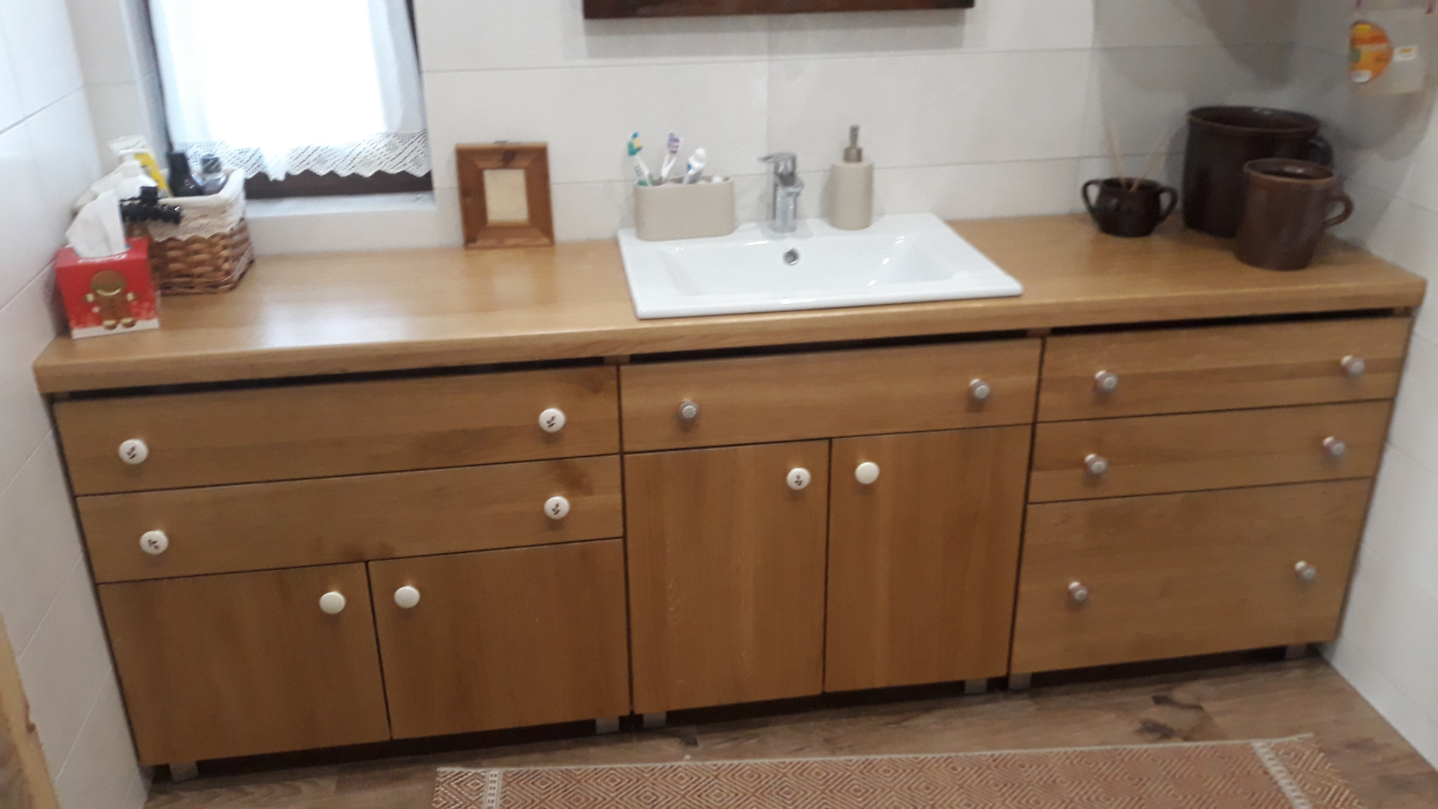 Drewniana szafka łazienkowa pod umywalką z białym blatem i okrągłymi uchwytami na tle białych płytek i okna z koronkową firanką.