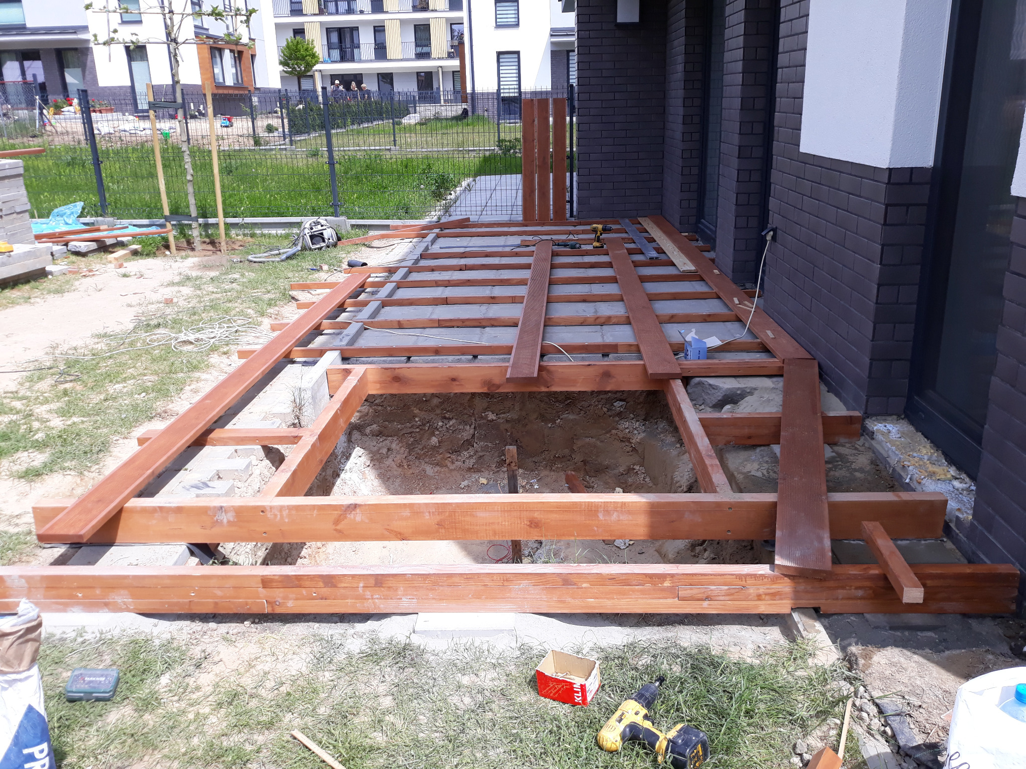 Konstrukcja drewniana tarasu w trakcie budowy, widoczne legary i deski, narzędzia pracy leżą na trawie, w tle budynek mieszkalny.