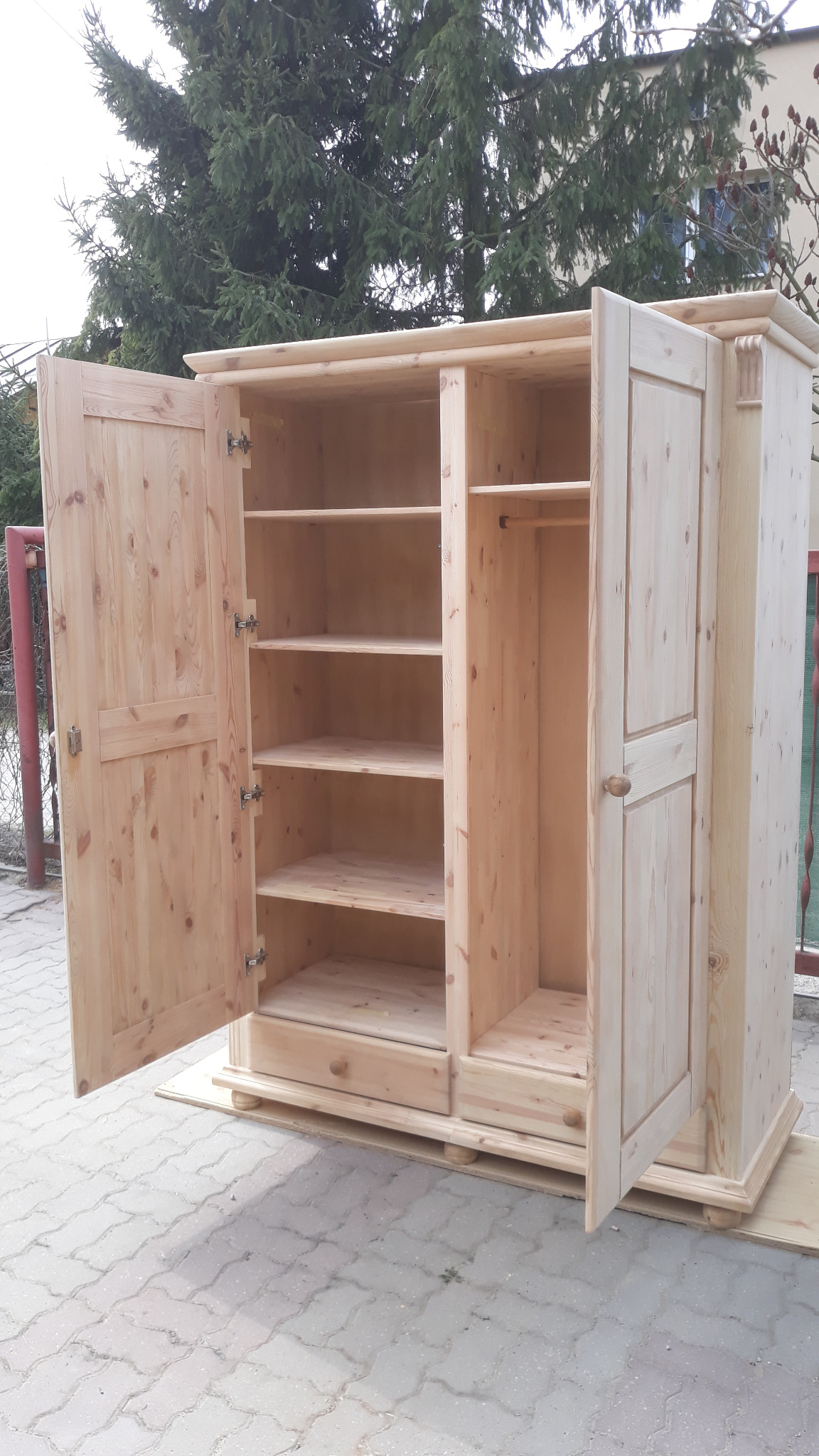 Surowa, drewniana szafa trzydrzwiowa z otwartymi drzwiami, ukazująca półki i drążek na ubrania, ustawiona na kostce brukowej przed zielonym drzewem iglastym.