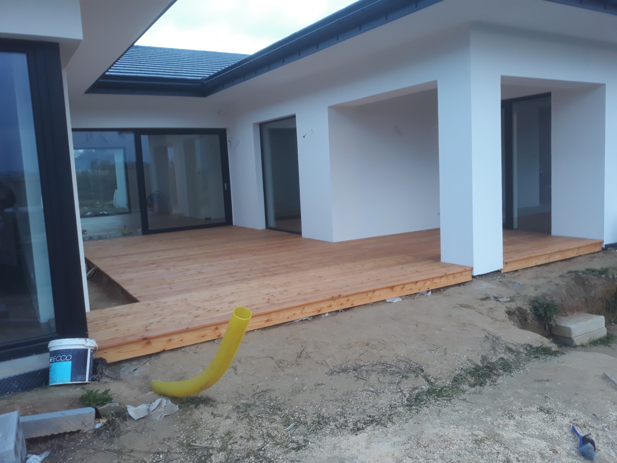 Nowoczesny taras drewniany z jasnego drewna przy białym budynku, widoczna żółta rura i wiadro z farbą na ziemi.