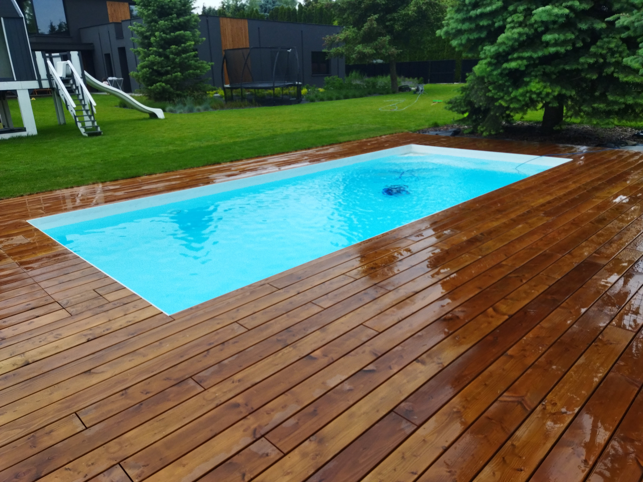 Prostokątny basen otoczony drewnianym tarasem z desek, widok na trawnik, nowoczesny dom z elementami drewna i zjeżdżalnią w tle.