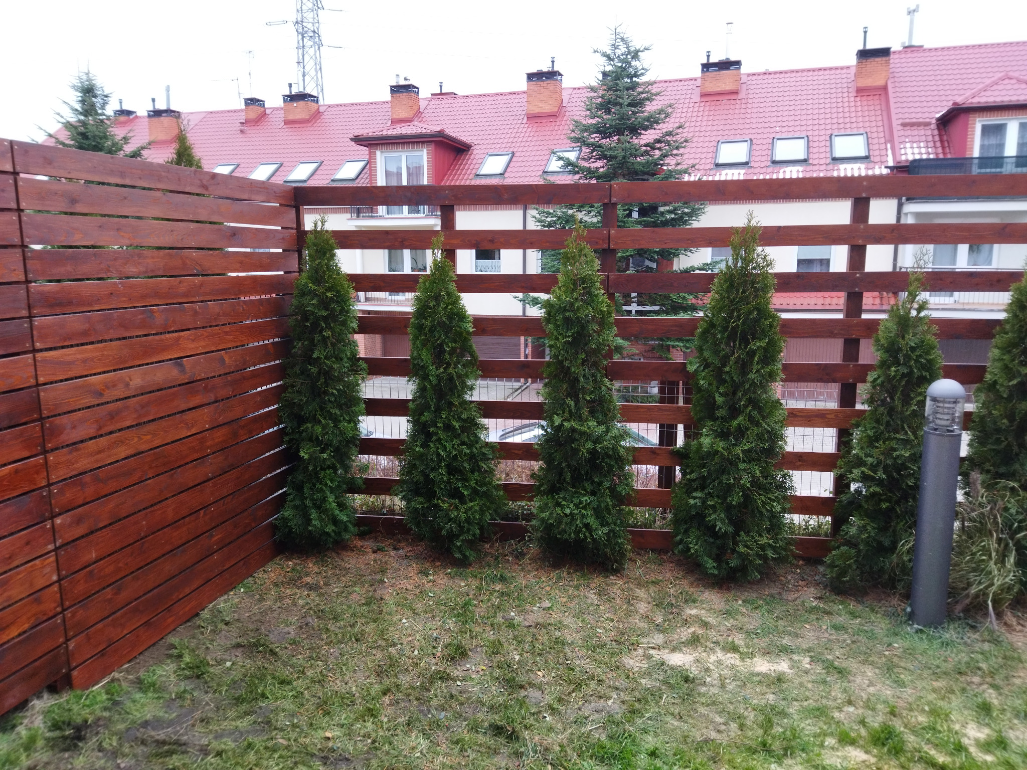 Nowoczesne ogrodzenie drewniane z poziomych desek, pomalowane na ciemny brąz, oddzielające posesję w Bełżycach; wzdłuż ogrodzenia posadzone tuje, w tle dom z czerwoną dachówką.