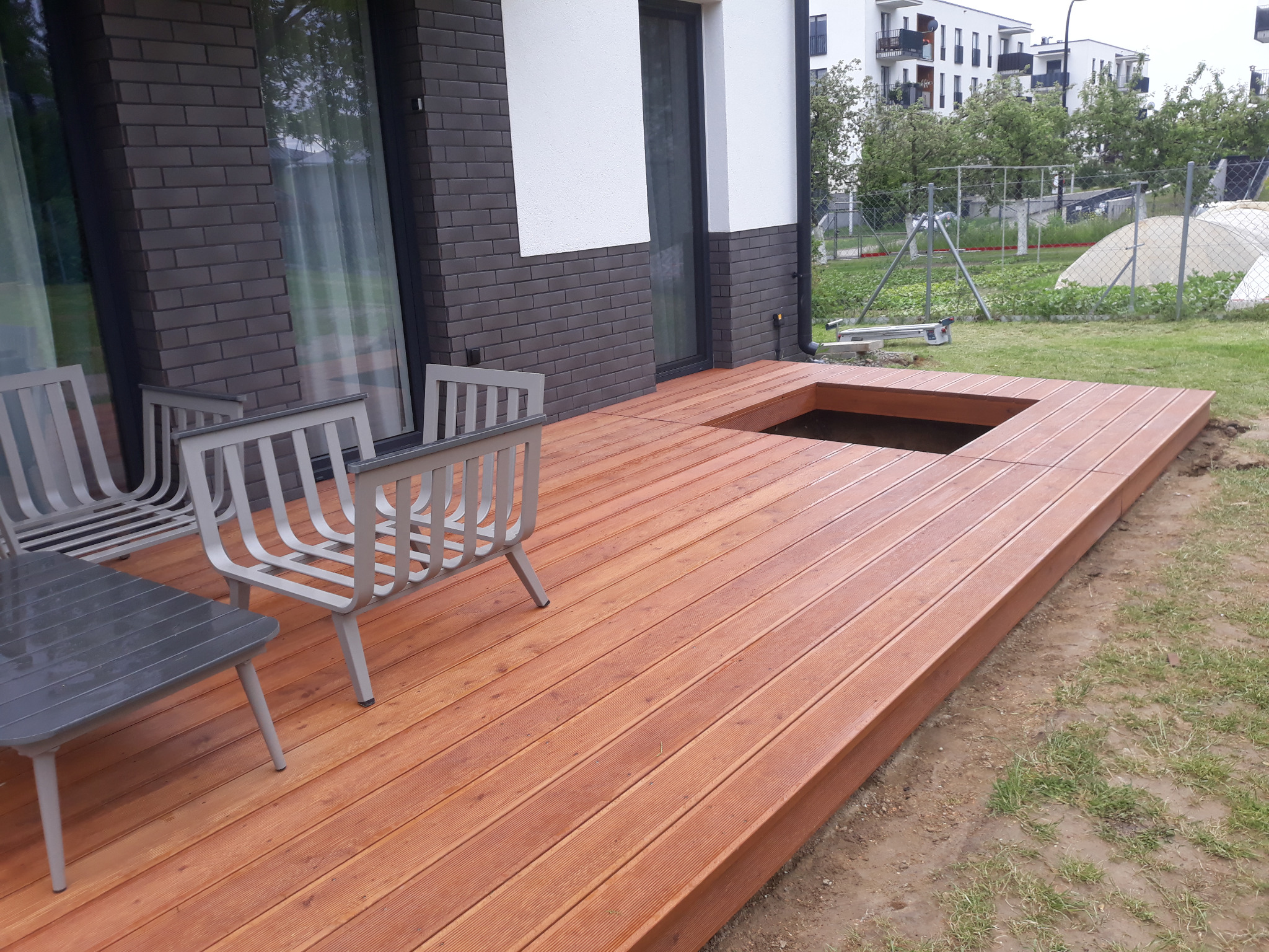 Nowoczesny taras z drewna egzotycznego z wycięciem na basen/oczko wodne, szare metalowe meble ogrodowe, dom w tle.