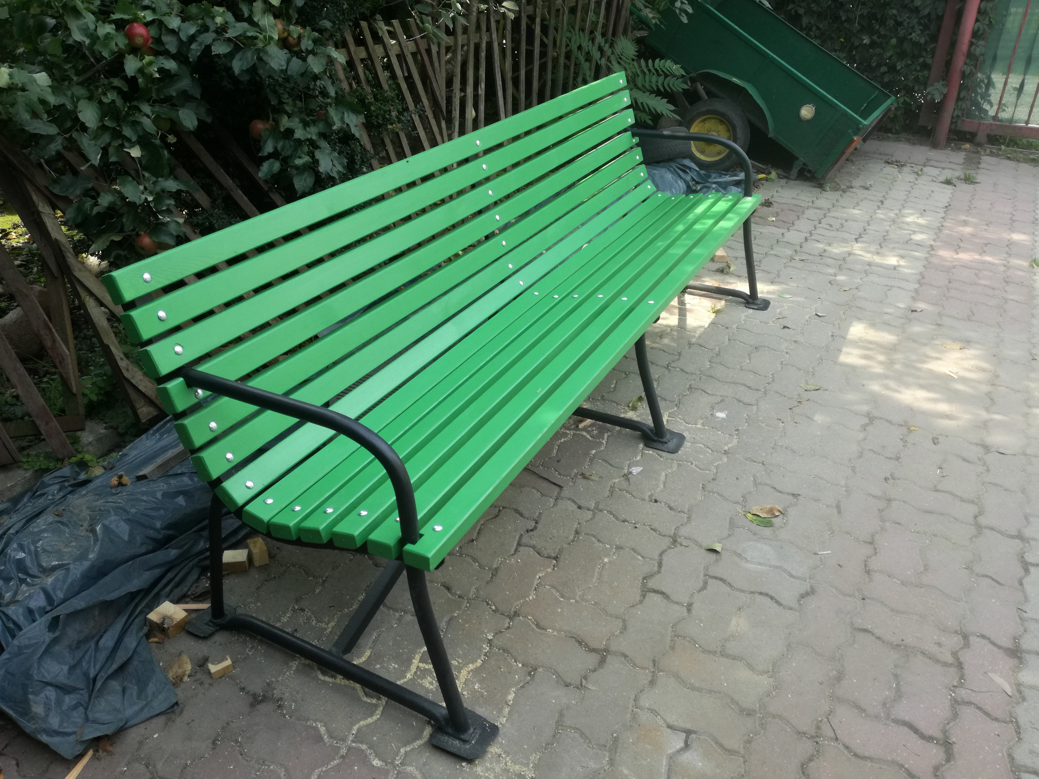 Świeżo pomalowana na zielono ławka parkowa z czarnymi metalowymi nóżkami, stojąca na kostce brukowej, z widoczną przyczepką w tle.