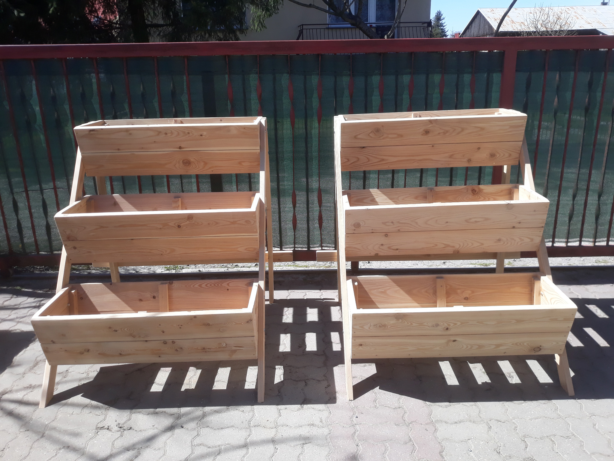 Dwie drewniane, trzypoziomowe donice ogrodowe ustawione na brukowanym tarasie w słoneczny dzień, z widocznym ogrodzeniem w tle.