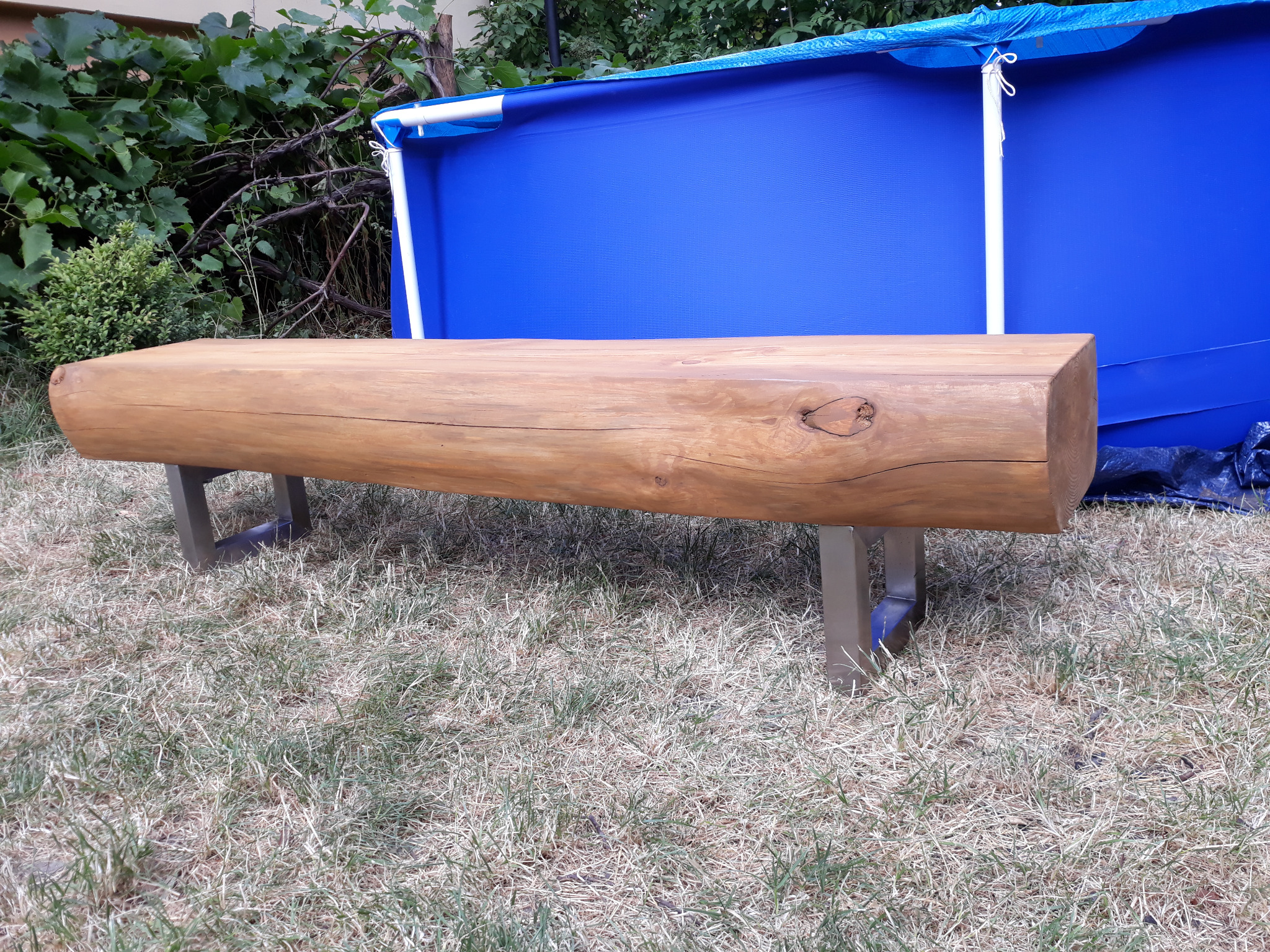 Ławka ogrodowa wykonana z grubego, naturalnego pnia drzewa z widocznym sękiem, wsparta na metalowych, kwadratowych nogach, na tle trawnika i częściowo widocznego, niebieskiego basenu ogrodowego.