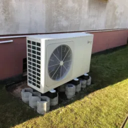 Zewnętrzna jednostka pompy ciepła Midea zamontowana na betonowych bloczkach na trawniku przy ścianie budynku.