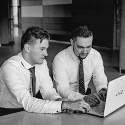 Dwóch mężczyzn w koszulach i krawatach pracuje wspólnie przy białym laptopie VAIO na jasnym stole w nowoczesnym wnętrzu z kolumnami, czarno-białe zdjęcie.